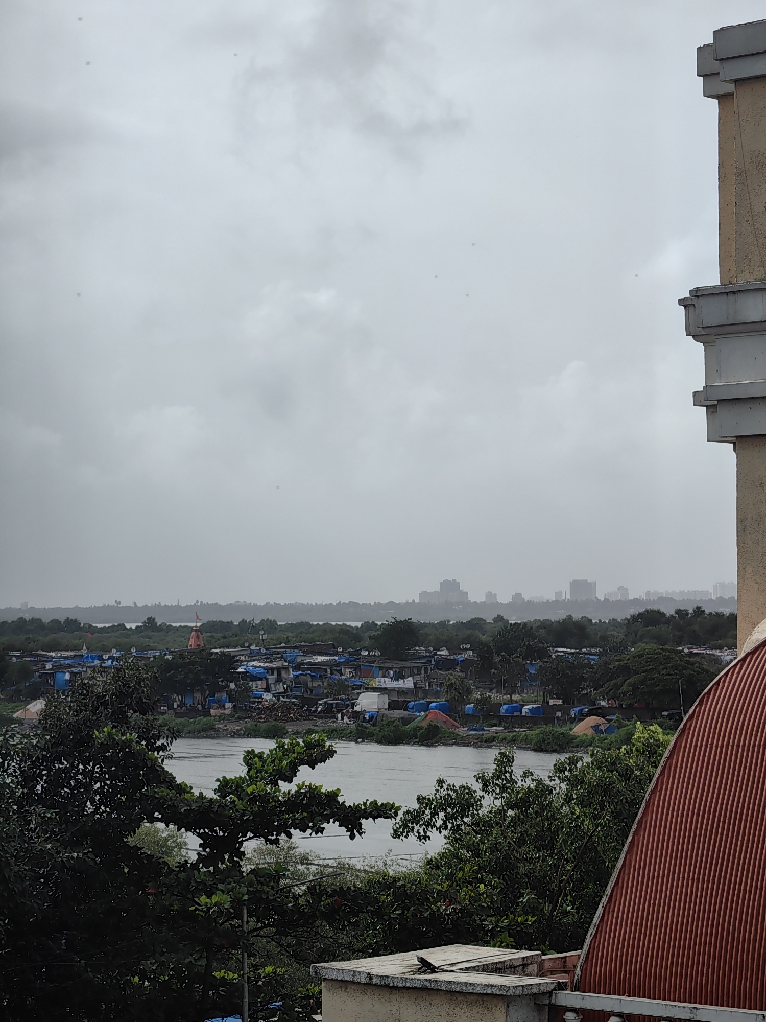 The image depicts an overcast view of a coastal area, likely in Mira-Bhayander, India, with a body of water in the mid-ground. Along the waterfront, there are numerous small dwellings with blue tarpaulin roofs, indicative of a slum or informal settlement. A prominent reddish-orange temple structure with a flag stands out amongst the houses. Trees and vegetation are scattered throughout the scene, bordering the water and the settlement. In the distance, a hazy urban skyline with tall buildings is visible. In the foreground, the edge of a building with a textured, corrugated red roof and beige concrete structure is present, with a small lizard visible on the concrete ledge. The sky is uniformly gray and cloudy, suggesting a dull or potentially rainy day.