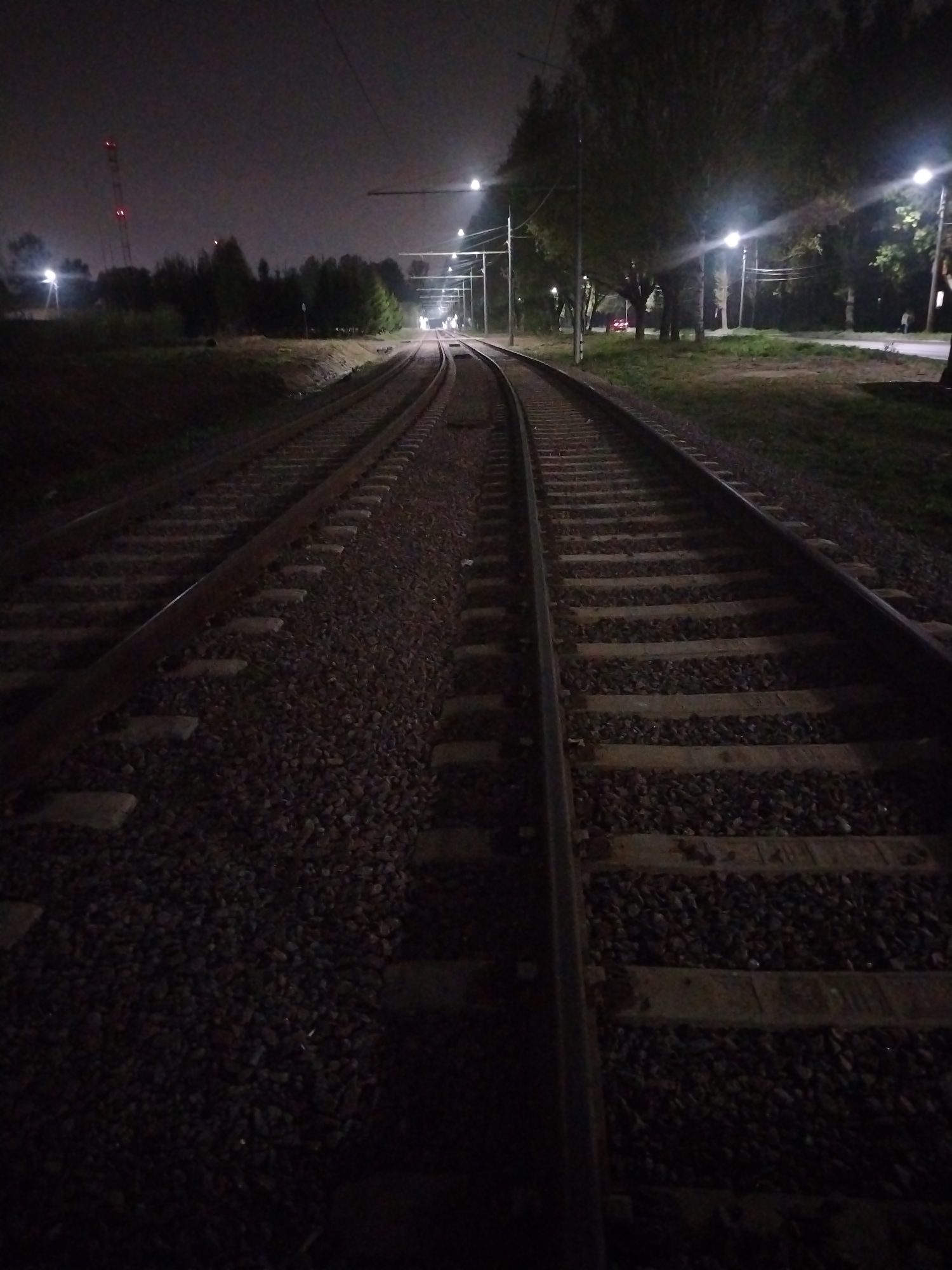 The image depicts a railway track at night, curving into the distance. The tracks are made of metal rails and wooden sleepers, with ballast consisting of small rocks filling the space between and around the sleepers. Streetlights illuminate the scene along the right side of the tracks, suggesting the presence of a road or path alongside the railway. On the left, there is vegetation, including trees and bushes. In the distance, a tower with red lights can be seen. Based on the scene's characteristics, it is likely taken in Yaroslavl, Russia where a train line exists near a park area. The scene is dark and quiet. There are no people visible in the frame.