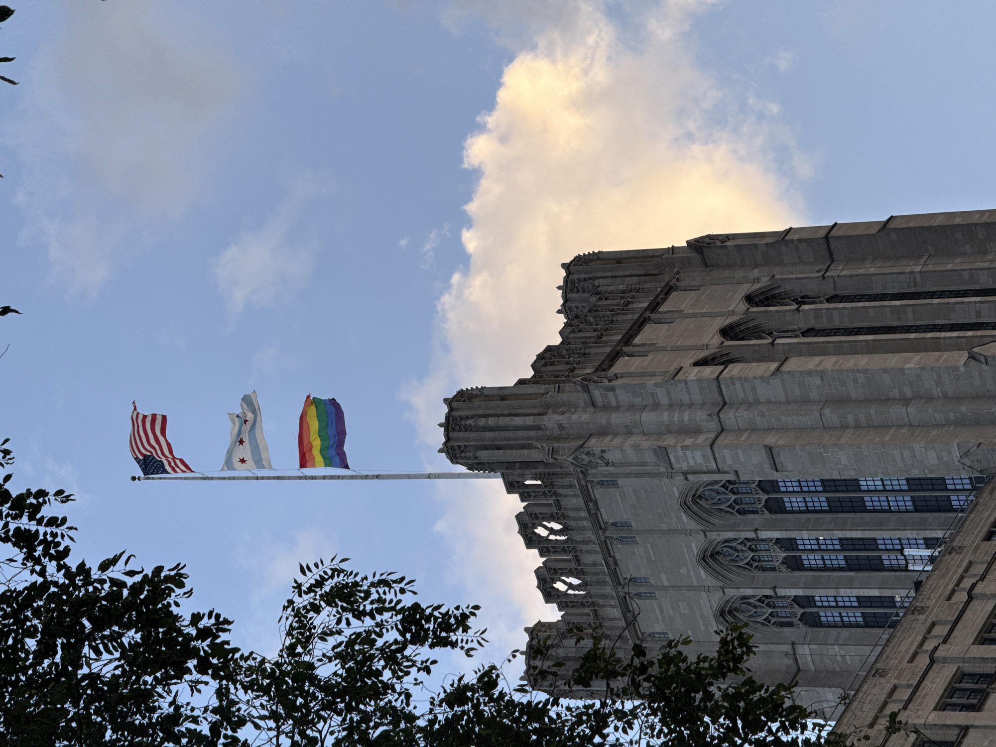 The image shows the top of a gothic-style building, likely the University of Chicago's Rockefeller Chapel, against a blue sky with scattered clouds. A flagpole stands to the right of the building, flying three flags: the American flag at the top, the Chicago city flag in the middle, and a rainbow pride flag at the bottom. Dark green leaves from a tree partially obscure the upper left portion of the image. The building's facade is made of light gray stone and features pointed arch windows with intricate tracery. The sky transitions to a lighter hue with hints of yellow and orange, possibly indicating sunset or sunrise. The overall impression is a serene and somewhat stately scene.