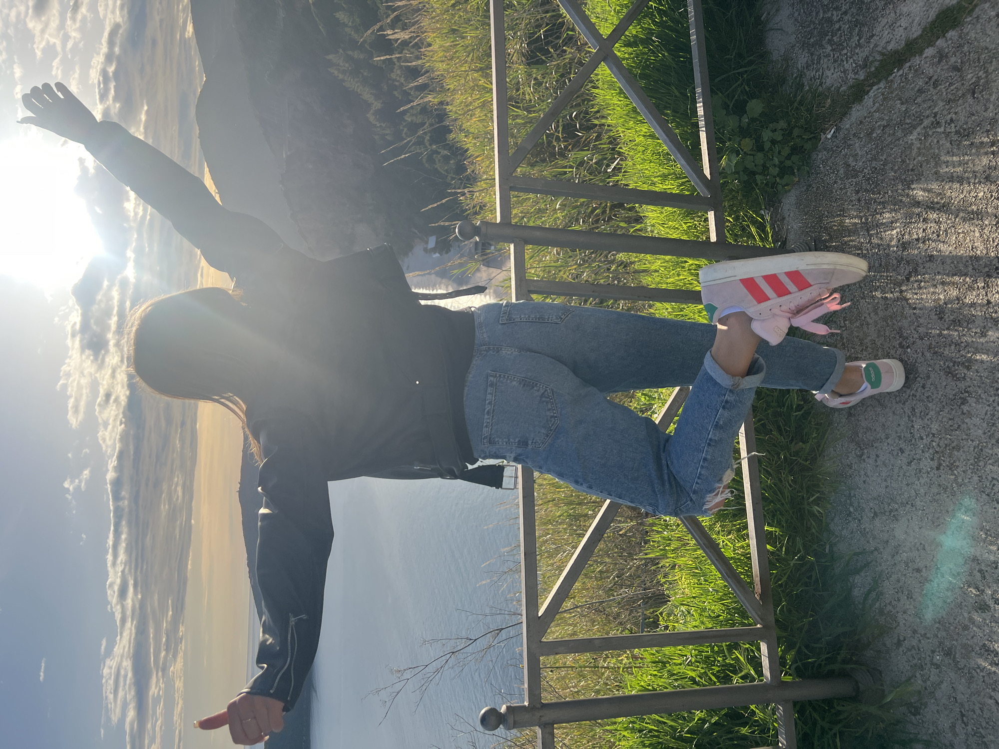 A person is standing on a paved surface next to a metal railing. The railing overlooks a body of water with a mountainous landscape in the background. The person has their arms outstretched. They are wearing a black jacket and light blue jeans. On their feet are pink and white Adidas sneakers. The environment includes grass and other vegetation. The location is Prijevor, Montenegro.