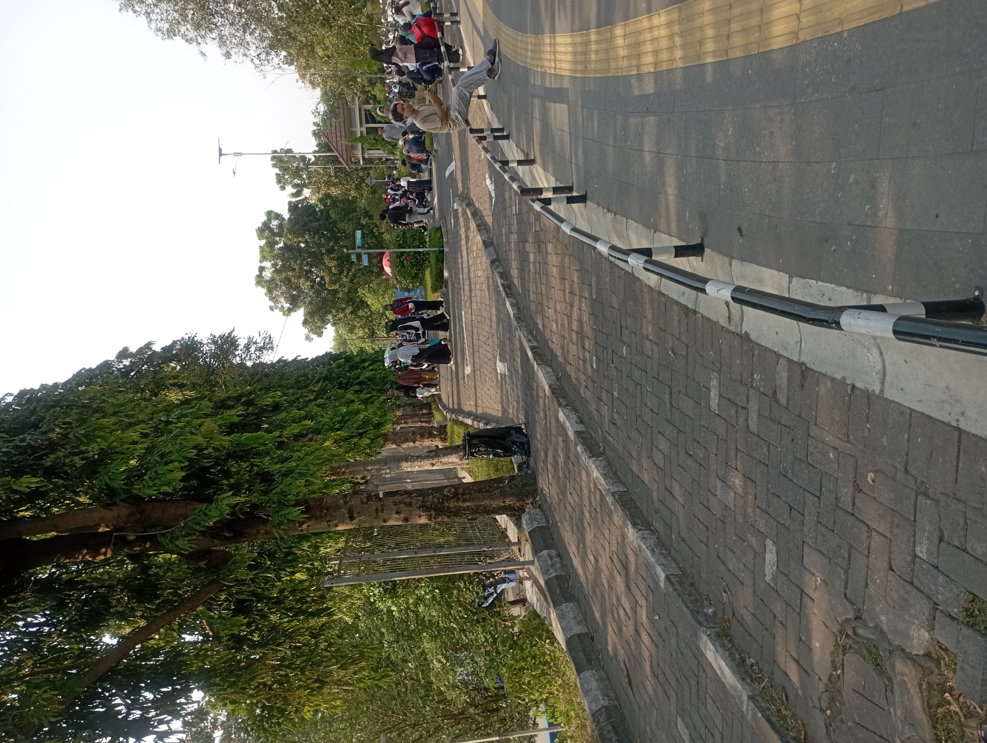 The image depicts an outdoor scene, likely in a park or public area in Depok, Indonesia, during the late afternoon as indicated by the warm, low-angled sunlight casting shadows. In the foreground, a paved walkway constructed with interlocking gray bricks curves to the right. A concrete curb separates the brick walkway from a sidewalk made of larger gray tiles, which is further bordered by a tactile paving strip of yellow tiles. A black and white striped metal railing runs along the edge of the sidewalk.
Several trees with lush green foliage are visible along the left side of the walkway, casting dappled shadows on the pavement. A tall, black trash bin with a plastic bag is situated near the base of one of these trees. Beyond the trees, a metal fence separates the walkway from a grassy area with some smaller trees.
In the middle ground and background, a diverse group of people are engaged in various activities. Some individuals are walking along the walkway, while others are sitting or standing in groups. Many are wearing traditional Islamic attire, such as headscarves and long dresses, suggesting a culturally significant gathering or a common practice in the area. One person, a young man wearing a light brown t-shirt and gray pants, is seated on the railing, looking at his phone. Further back, a small, open-air pavilion with a reddish-brown roof is visible amidst the trees, and a street lamp with solar panels stands tall. The overall atmosphere appears to be one of casual social interaction and relaxation in a well-maintained urban green space.