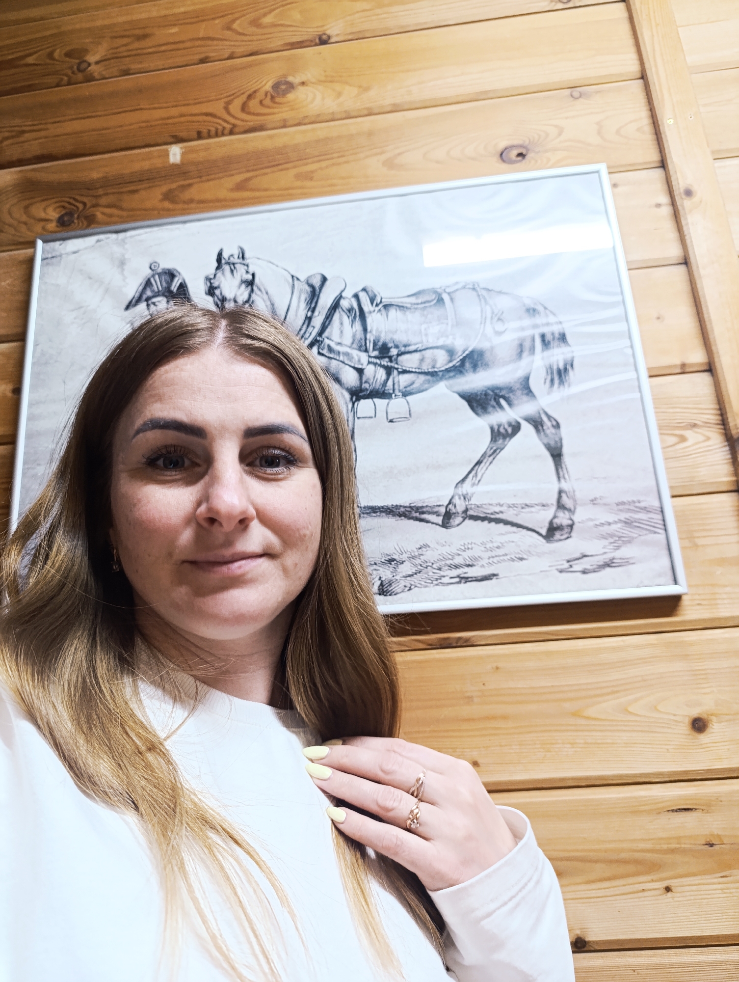 A woman is centered in the frame, with a horse-themed picture behind her. The location is a room with wooden walls. The woman has fair skin, brown hair, and is wearing a white top. She is wearing rings on her fingers and has light yellow nail polish. The picture behind her is framed and depicts a horse with a rider. The wooden walls have a rustic appearance.