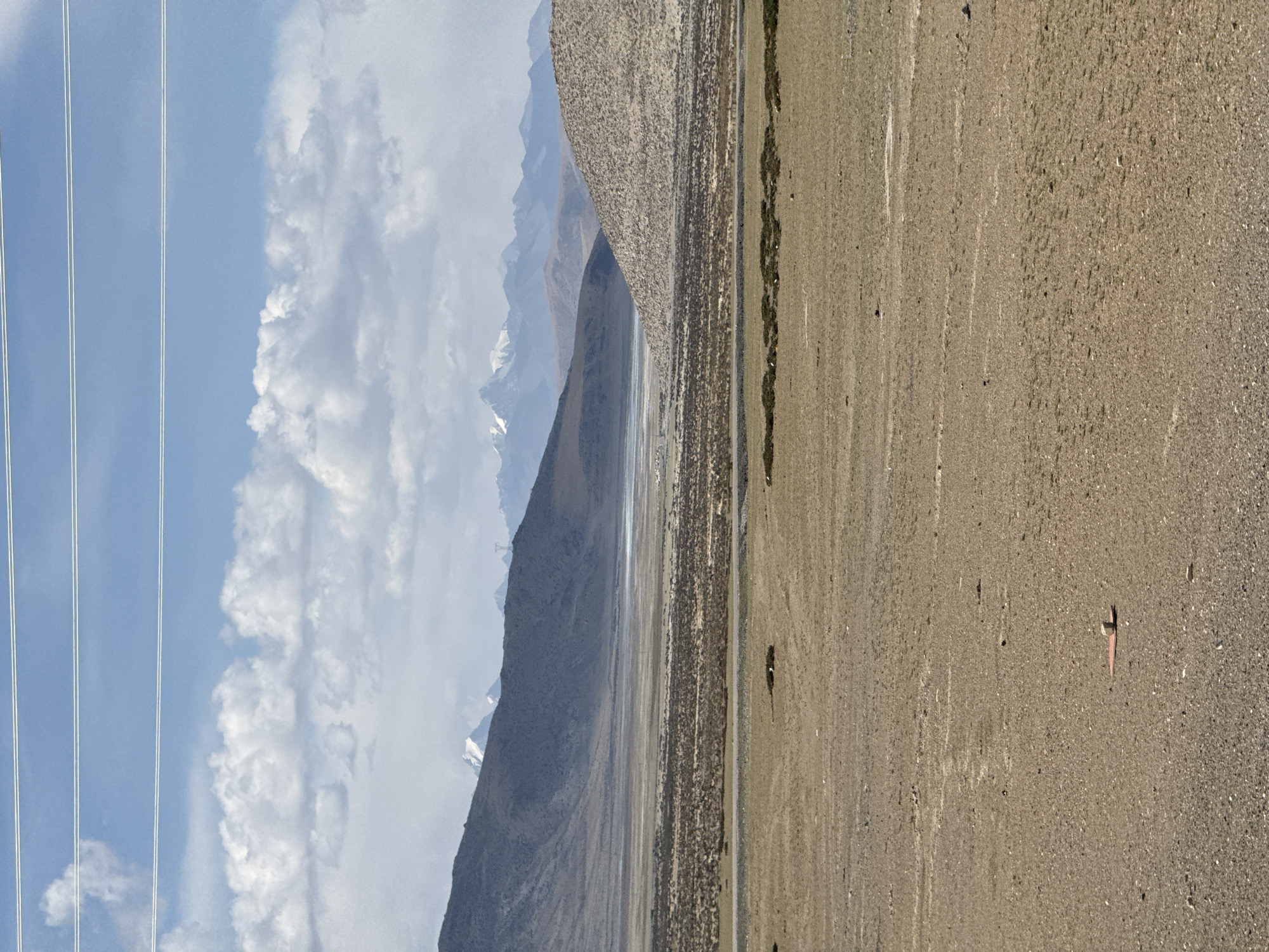 The image depicts a landscape in Gyirong County, China. The sky is partly cloudy with power lines visible. Mountains are in the background with snow-capped peaks. A dry, gravelly plain stretches across the foreground. A road is between the plain and mountains. Sparse vegetation grows along the roadside. A small, red object is on the plain.