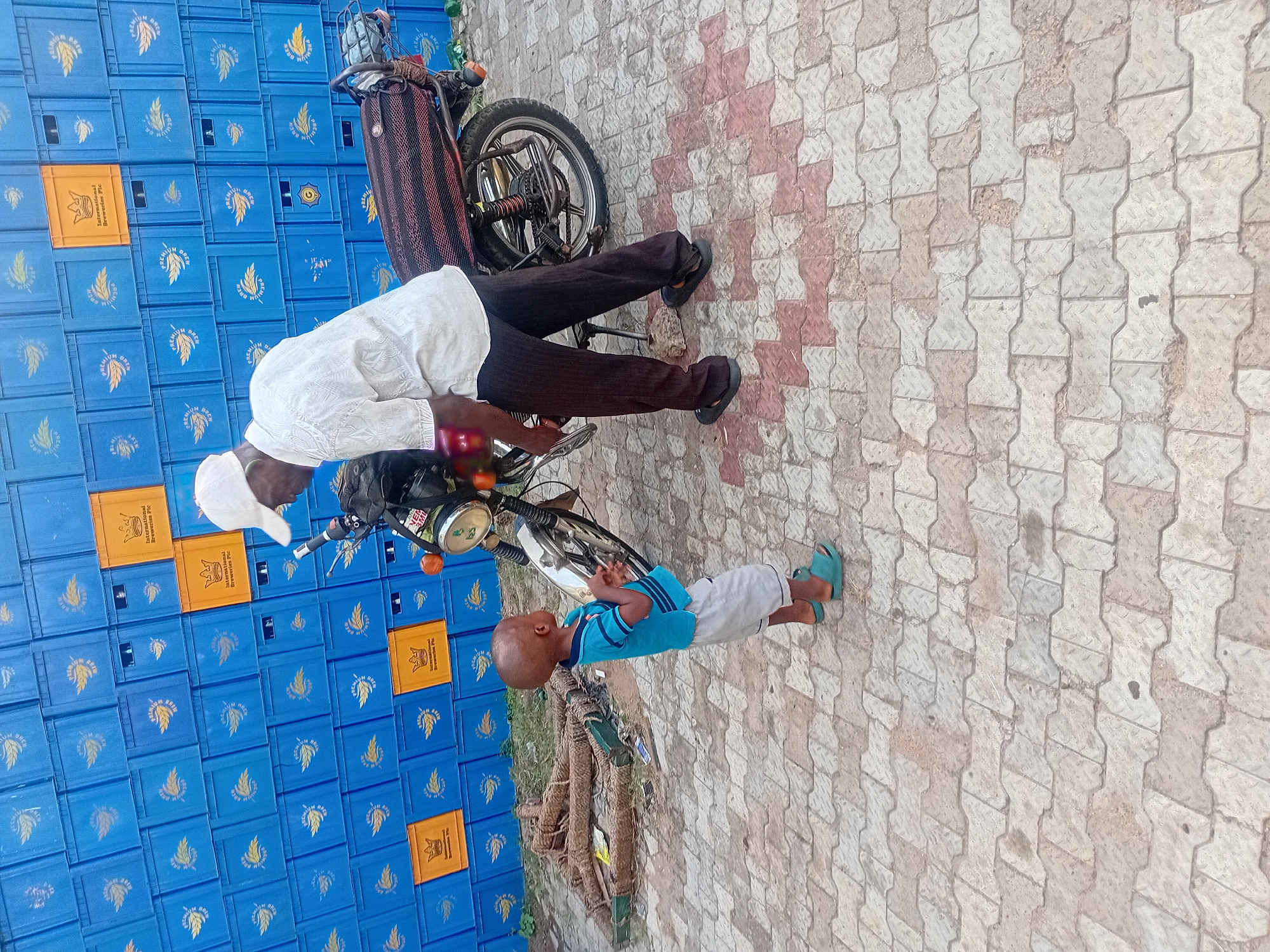 This outdoor image, captured in Lokoja, Nigeria, features an adult male, a young child, and a motorcycle set against a backdrop of stacked beverage crates. The adult, dressed in a white long-sleeved shirt, dark trousers, and a white cap, is leaning over a dark-colored motorcycle, seemingly engaged in an activity involving the vehicle. The motorcycle has chrome components and visible orange turn signals.
A young child, appearing to be male, with dark skin and a bald head, stands nearby, observing the adult or the motorcycle. He is wearing a blue and white striped t-shirt, light-colored shorts, and teal sandals.
The background is dominated by a tall stack of blue plastic crates, with several orange crates interspersed among them. The blue crates are marked with "PREMIUM BREW" and a logo depicting a wheat stalk or grain. The orange crates display "International Breweries PLC" and a crown-like emblem, indicating their use in the beverage industry.
The ground is paved with interlocking concrete blocks, primarily in light grey and beige tones, with some reddish-brown sections forming a pattern. Coiled ropes or thick wires are visible on the ground next to the crates. The scene is well-lit, suggesting it is daytime, portraying a calm, everyday moment in what appears to be a commercial or storage area.