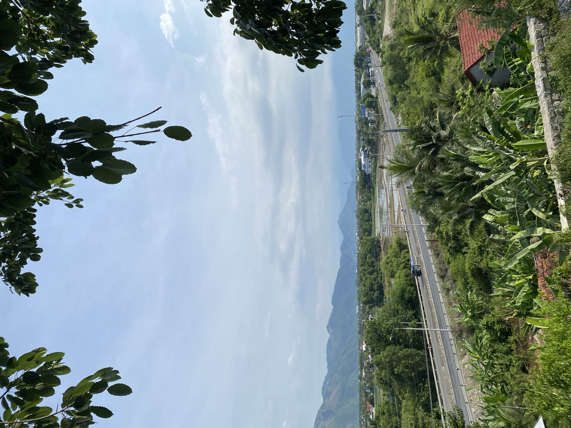 The image depicts a wide, high-angle view of a landscape in Tay Nha Trang Ward, Vietnam, on a partly cloudy day. In the foreground, lush green foliage, including banana trees and other tropical vegetation, dominates the bottom half of the frame, with hints of tiled rooftops visible. A low stone wall runs along the very bottom edge. A highway, with two lanes of traffic, cuts horizontally across the middle ground, with a single vehicle, a dark-colored truck, visible on the left lane heading away from the viewer. To the left and right of the highway are expanses of green trees and some scattered buildings. In the background, a range of hazy blue mountains forms the horizon, with a clear sky above, broken by streaks of white clouds. The overall impression is one of a verdant, semi-rural environment transitioning towards development.