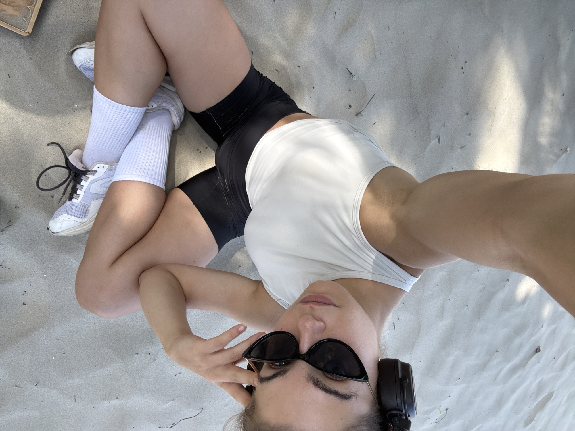 A young woman lies on a sandy surface, likely a beach, wearing black athletic shorts, a white tank top, white athletic socks, and light-colored sneakers. She is also wearing dark sunglasses and headphones. The scene is lit with natural light. There is no visible evidence to conclusively place the location in Đà Nẵng, Vietnam.