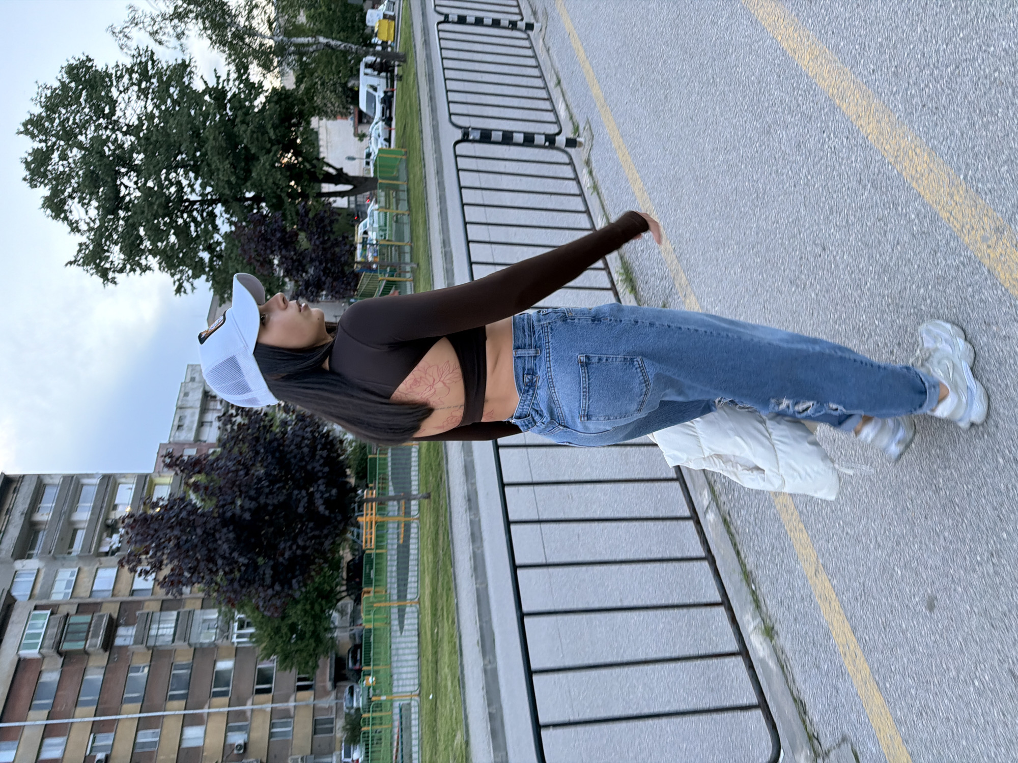 A woman is standing on a street in what appears to be an urban setting. She is wearing a white cap, a long-sleeved brown shirt that exposes her midriff, blue jeans, and light-colored sneakers. A white jacket is draped over her arm.
In the background, there are buildings, trees, and a fence. A street with yellow lines is visible on the ground. Based on the buildings and environment, it is possible this is a photograph taken in the city of Gabrovo, Bulgaria.