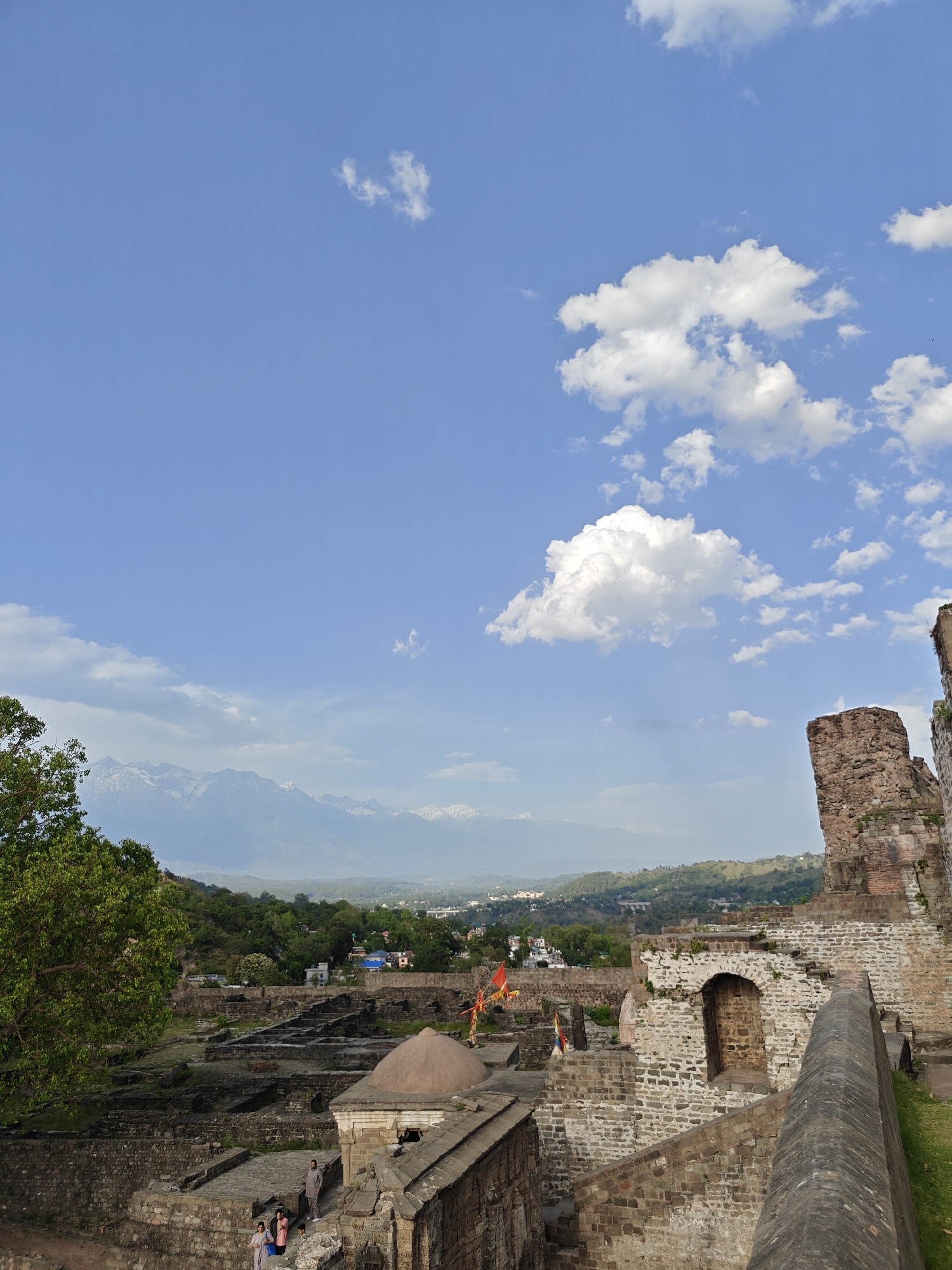 The image shows the Kangra Fort in Kaswara, India. The foreground features the weathered stone ruins of the fort, with a domed structure visible among the remains. A small group of people, including women and children, are walking on the stone structure. Beyond the fort, a green valley stretches out towards a mountain range with snow-capped peaks. The sky is blue with scattered white clouds.