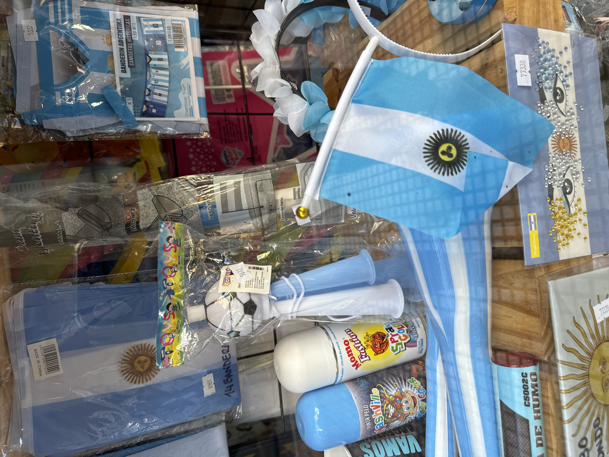 A close-up of a display of Argentina-themed merchandise, likely for a celebration of the Argentinian national team. Items include:
* Multiple Argentinian flags in varying sizes.
* Headbands with blue and white floral decorations.
* Horns, spray paint, and smoke effects in Argentinian colors.
* Packaged banners and historical jersey-themed items.
* Adhesive rhinestones and embellishments for DIY projects.
* Product packaging in Spanish.
The scene is brightly lit, suggesting indoor lighting or natural light from a window. The setting appears to be a retail display, possibly in a store or souvenir shop. The environment seems crowded, with items packed together closely on a shelf. The image is focused and close, capturing the details of the merchandise.