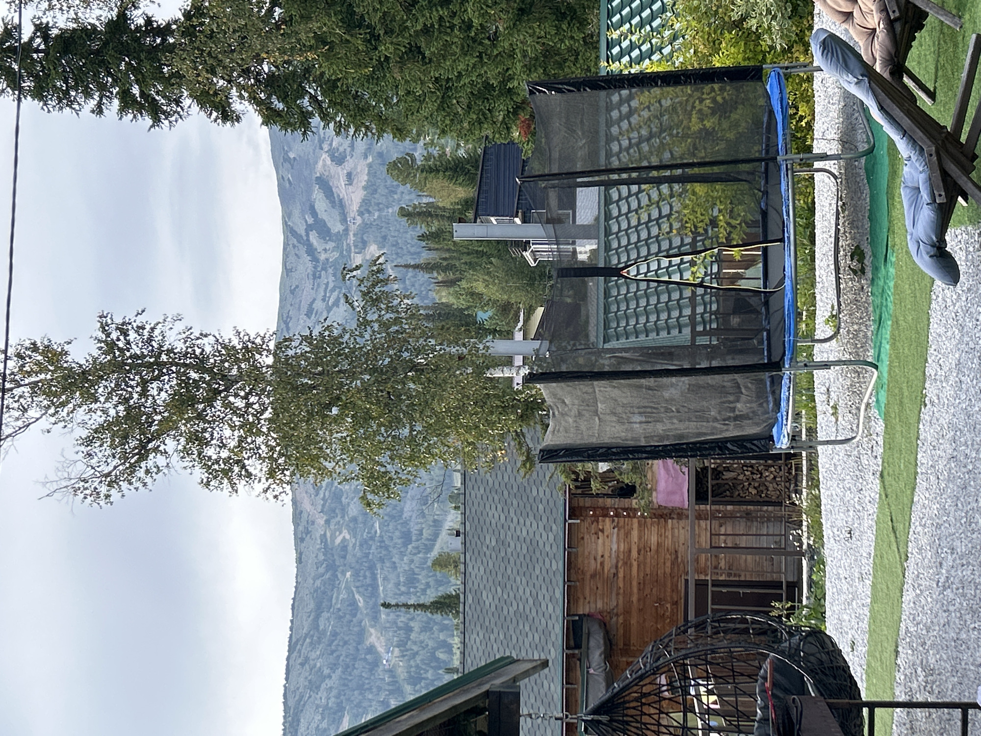 The image depicts a backyard scene in Sheregesh, Russia, with a backdrop of rolling, forested mountains under a cloudy sky. In the foreground, a round black hanging chair is visible on the left, followed by a trampoline with a black safety net and a blue jumping mat. A grey wooden lounge chair with a blue cushion is in the lower right corner, and part of another brown cushioned lounge chair is to its right. The ground is covered with light grey gravel and patches of artificial green turf.
Behind the trampoline, a wooden cabin with a dark green shingled roof is partially visible. Further back, there are more buildings with dark green roofs and a tall, slender evergreen tree. The midground and background are dominated by dense coniferous forests covering the mountains, with some evidence of ski slopes or trails visible on the distant slopes. The overall atmosphere is serene and suggests a mountainous resort or private residence setting.