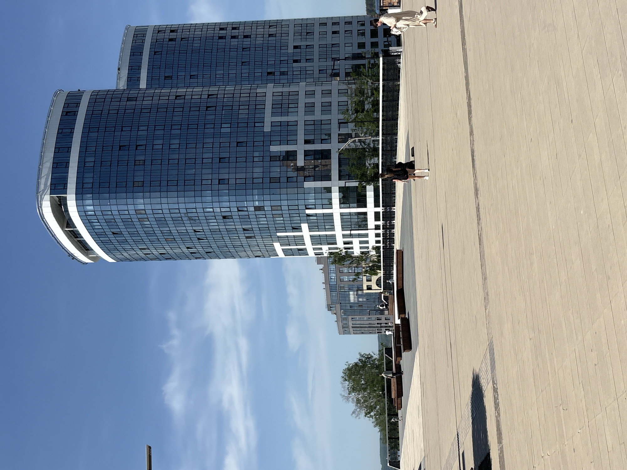 The image depicts a public square in Izhevsk, Russia, featuring modern architecture and pedestrian activity. In the background, two conjoined high-rise buildings dominate the skyline with curved facades, clad in blue-tinted glass windows arranged in a grid. A white, grid-like pattern extends across the lower section of the facade, adding a design element. The sky is a clear blue with wispy white clouds.
Two women are visible in the foreground. One, wearing a short black dress and white sneakers, is walking along the paved ground of the square. The other woman, dressed in a long beige dress, is also walking.
Benches line the perimeter of the square. Lampposts stand along the walkway. In the distance, trees provide greenery to the scene.
