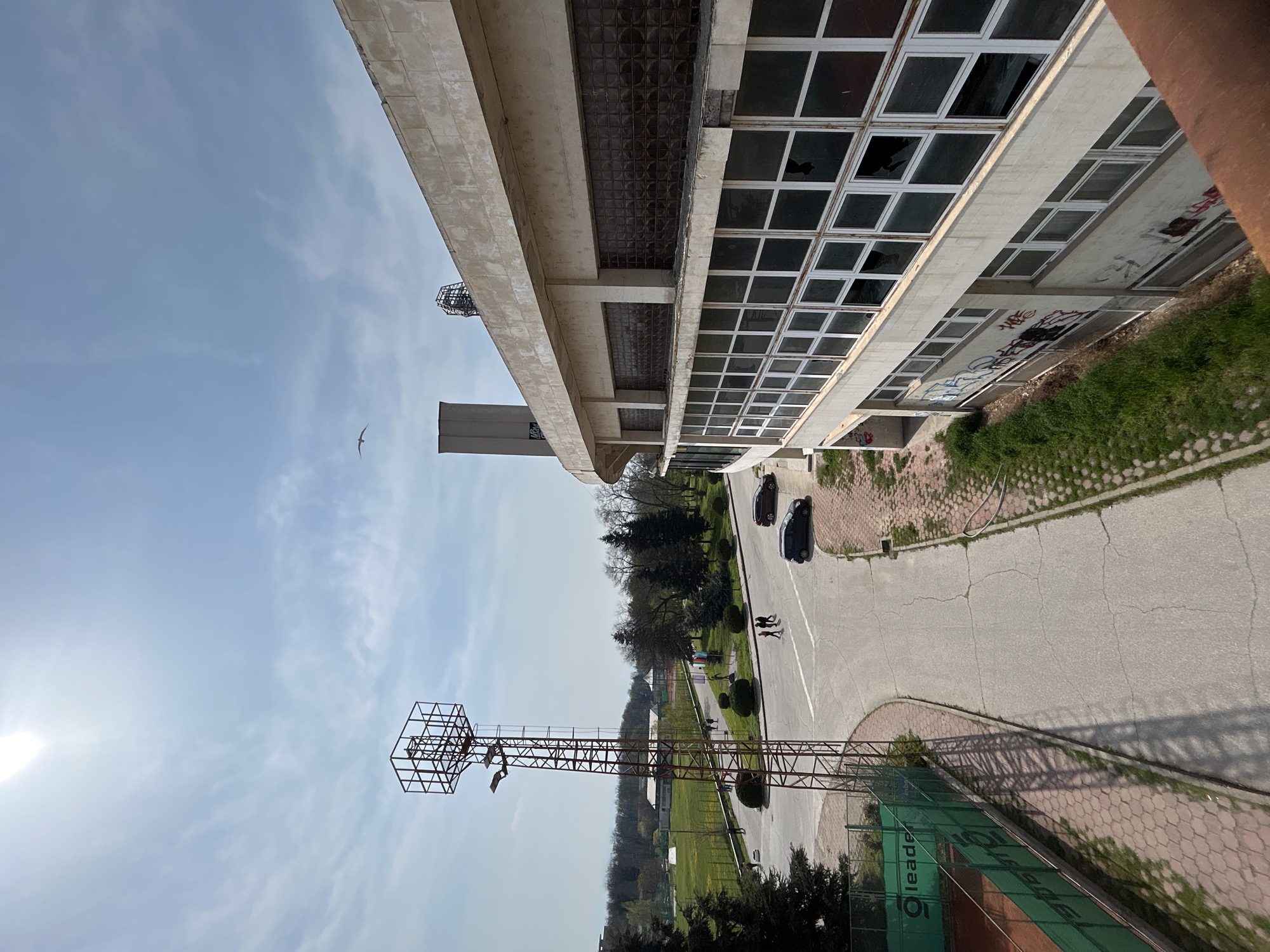 The image depicts an elevated view overlooking a street and park area in Plovdiv, Bulgaria. A large, weathered building dominates the upper portion of the frame, featuring multiple rows of windows, some broken. Graffiti is visible on the building's lower exterior walls.
Below the building, a two-lane road curves through the scene, with two cars parked along the roadside. Two individuals with a dog are walking on the sidewalk.
In the foreground, a rusted metal structure, likely a lighting tower for a sports field, extends upwards. A tennis court with a green windscreen is visible beneath the tower. Trees and grass cover the park area beyond the road.
In the sky, there is a bird and a tall tower.