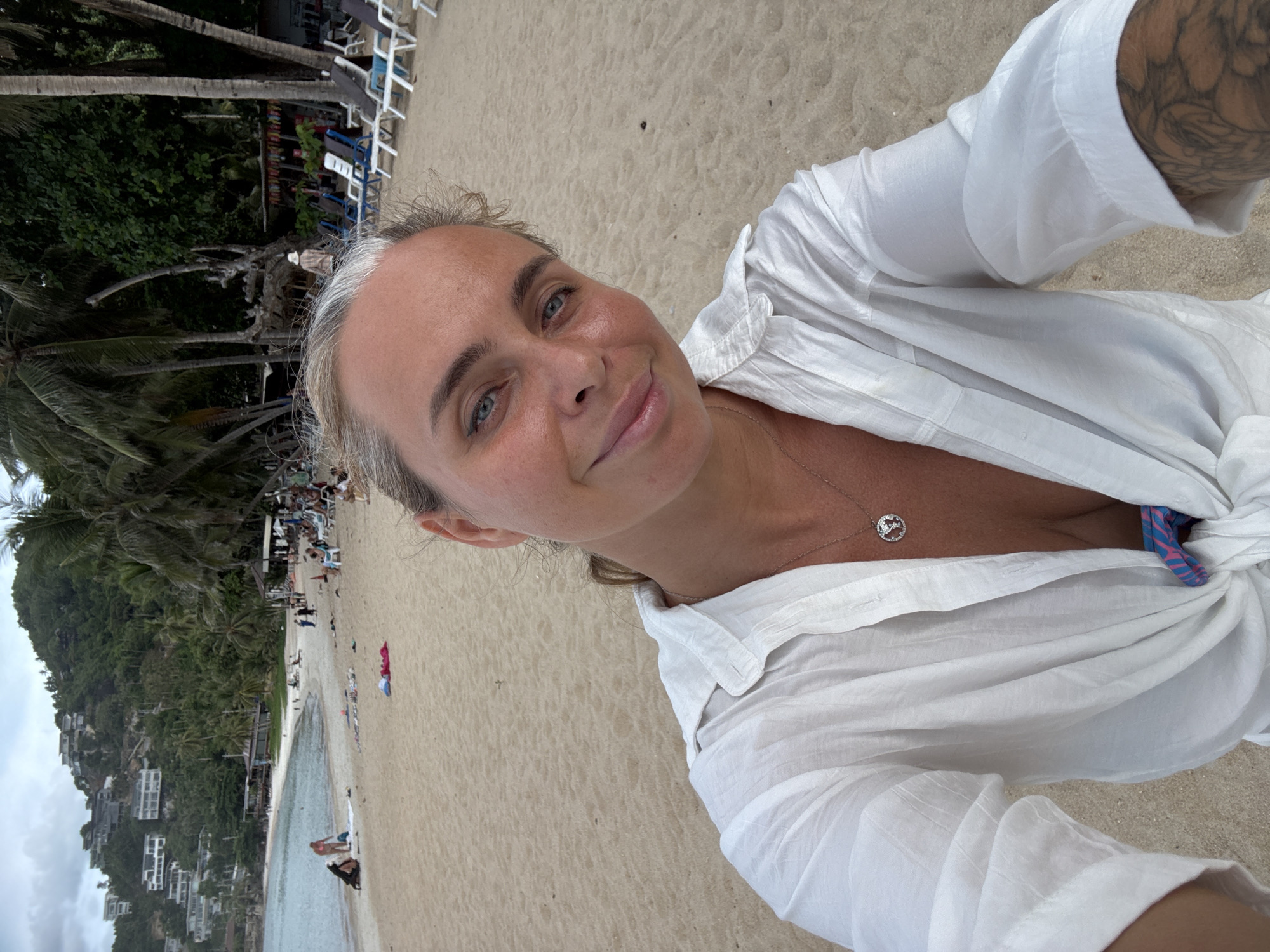 In the city of Baan Chaweng Noi, Thailand, a woman with blonde hair is taking a selfie on a sandy beach. She is wearing a white unbuttoned shirt tied at the waist, revealing a patterned bikini top. A necklace with a globe pendant adorns her neck. Her left arm shows a visible tattoo. The beach behind her is populated with a few people, lounge chairs, and palm trees. In the background, buildings are visible on a lush green hillside overlooking the ocean. The sky is overcast.