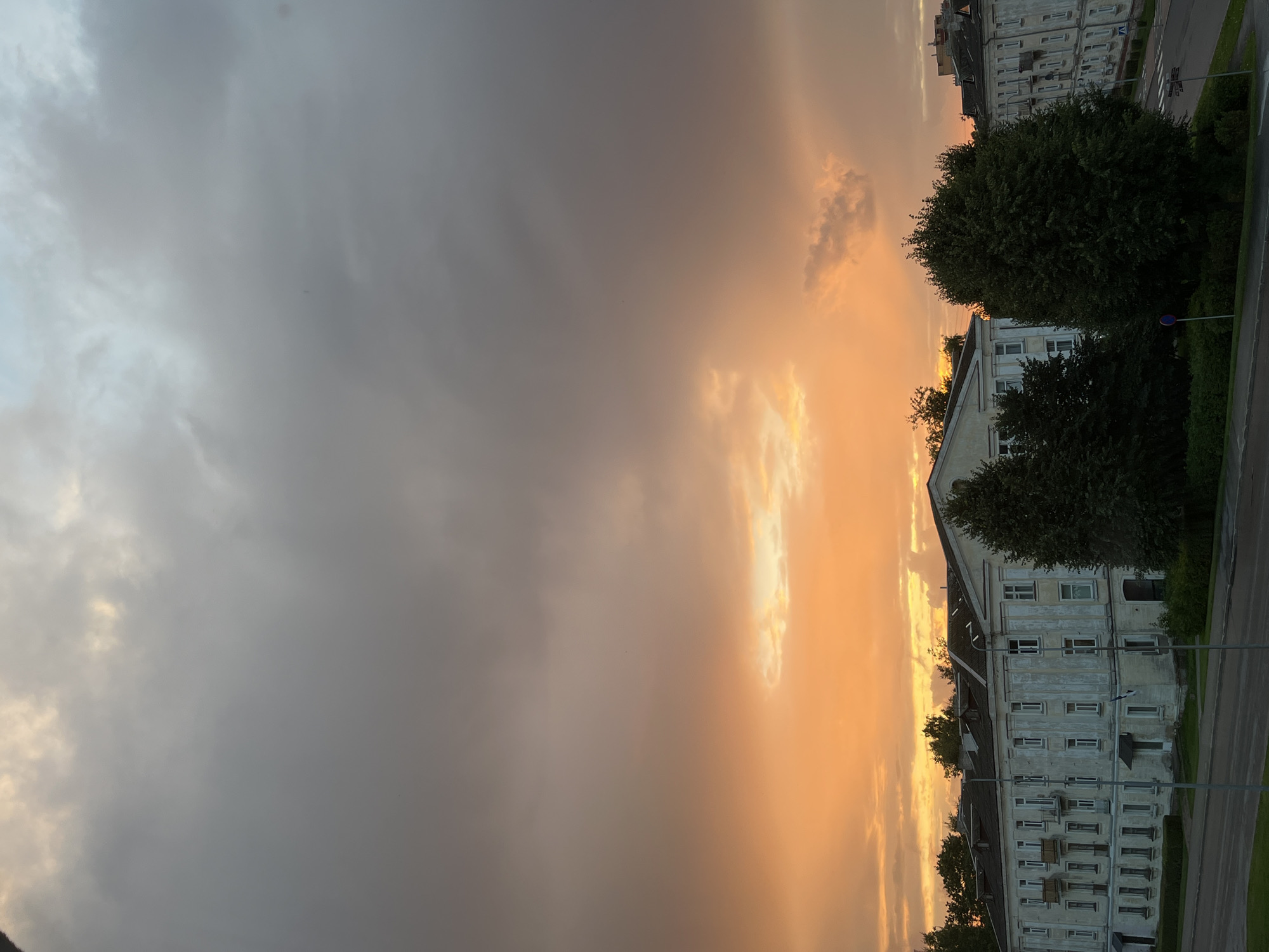 The image shows a sunset over the city of Kohtla-Järve linn, Estonia. The sky is filled with dramatic clouds in shades of orange, yellow, and gray. In the foreground, there are several Soviet-era apartment buildings, with their characteristic pale facades and numerous windows. Lush green trees partially obscure the buildings, and a paved road runs in front of them, with some streetlights visible. The overall impression is one of a quiet, somewhat somber urban landscape under a powerful sky.