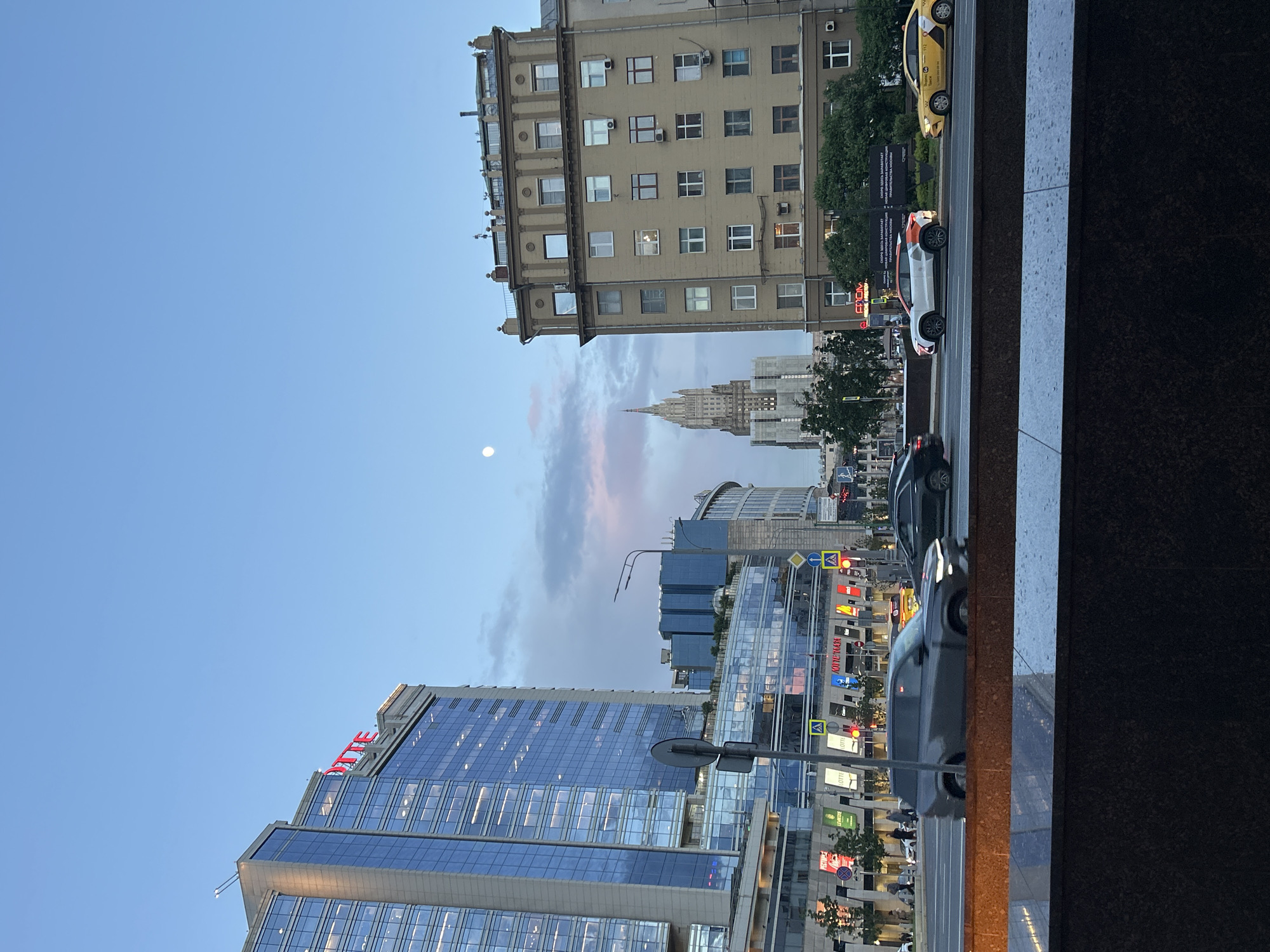 The image shows a street scene in Moscow, Russia. In the foreground, there's a dark granite or marble wall. The street is visible with cars driving on it, including taxis. Several buildings are in view. On the left, a modern glass skyscraper with the red word "LOTTE" on top. In the distance, a Stalinist skyscraper is visible, possibly one of the Seven Sisters. The sky is blue with some clouds. There is a small full moon. The image is rotated 90 degrees clockwise.