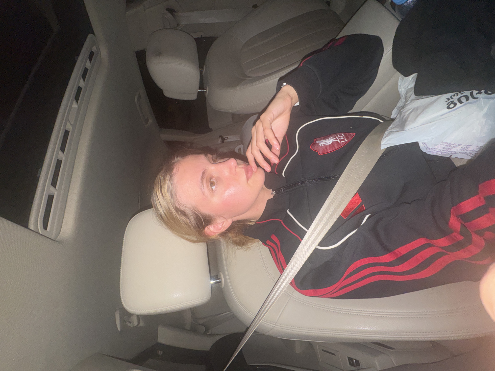 The image shows a young individual, likely female, lying horizontally across the back seats of a car at what appears to be night. They are wearing a black track jacket with red stripes on the sleeves and white piping. A red and white logo, resembling the Liverpool Football Club crest with "LFC", is visible on the jacket's chest. A light-colored seatbelt is fastened across their body.
The car's interior features light beige/tan leather seats and headrests. The windows show darkness, indicating nighttime. A white plastic bag rests near the person's feet, displaying visible text in Georgian script, including the word "გაიგე" (meaning "understand" or "find out") and the number "3000". The person looks towards the camera with a neutral expression, one hand near their chin. The scene is set inside a car in Tbilisi, Georgia.