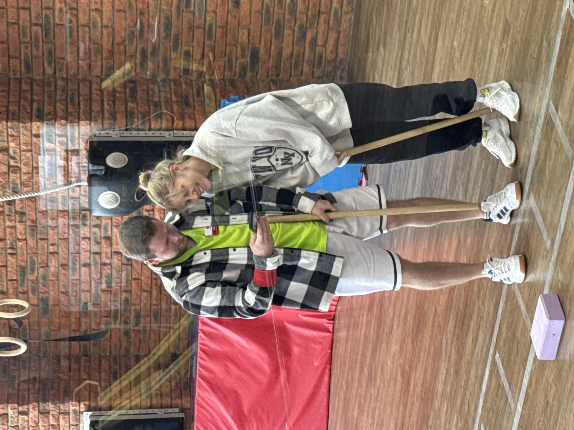 The image shows two people indoors, apparently in a gym setting. The foreground features a red mat near a wooden floor with white line markings. To the right is a purple block. A man in a bright green shirt, white shorts with black trim, a black and white checkered shirt, and white sneakers stands holding a wooden pole. Next to him, a woman in a gray sweatshirt with a logo, black pants, and white sneakers also holds the pole. They appear to be interacting or coordinating an exercise. In the background, there's a brick wall and some equipment mounted on it. Based on the presence of a gym setting with particular equipment and clothing, it is likely that this photograph was taken at the Chaikovsky, Russia, gym facility.