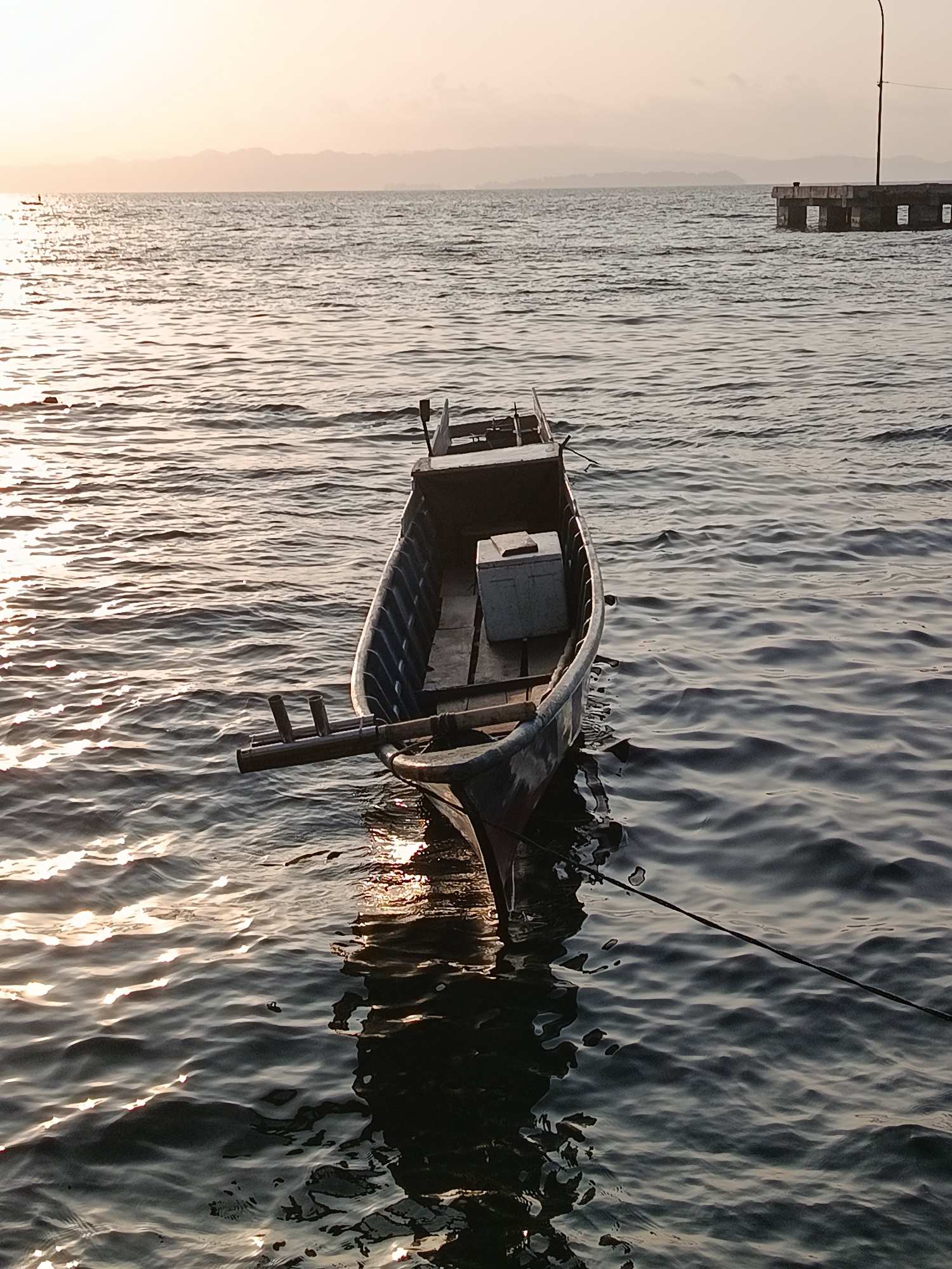 An empty, weathered fishing boat is moored in the tranquil waters of Mansalean, Indonesia, at dusk. The boat, with its faded blue hull and worn wooden interior, gently bobs on the rippling surface of the sea. A gray cooler is visible inside the boat. In the distance, the hazy silhouette of mountains frames the horizon, and a concrete pier juts out into the water, a solitary post with a light fixture marking its end. The setting sun casts a warm, golden glow across the water, creating shimmering reflections that dance on the waves. The scene is one of peaceful solitude, a quiet moment captured as day transitions into night in the coastal town.