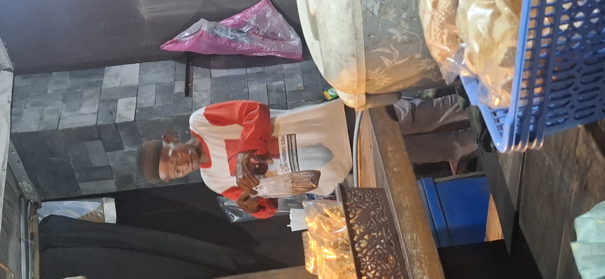 An elderly man, wearing a brown cap and a long-sleeved shirt with white and red sections, stands in front of a stall. He is holding a clear plastic bag containing a dark substance. To his left, on a table, are several plastic-wrapped snacks in a woven basket. To his right, a large, light-colored container with floral patterns is visible, and in the foreground, a blue plastic basket holds more plastic-wrapped snacks. The background features a dark wall with a section covered in dark grey rectangular stones. A pink plastic bag hangs on the wall to the man's right. The scene appears to be an outdoor stall, likely in a marketplace or street-side food vendor setting, possibly in Colomadu, Indonesia, given the context.
