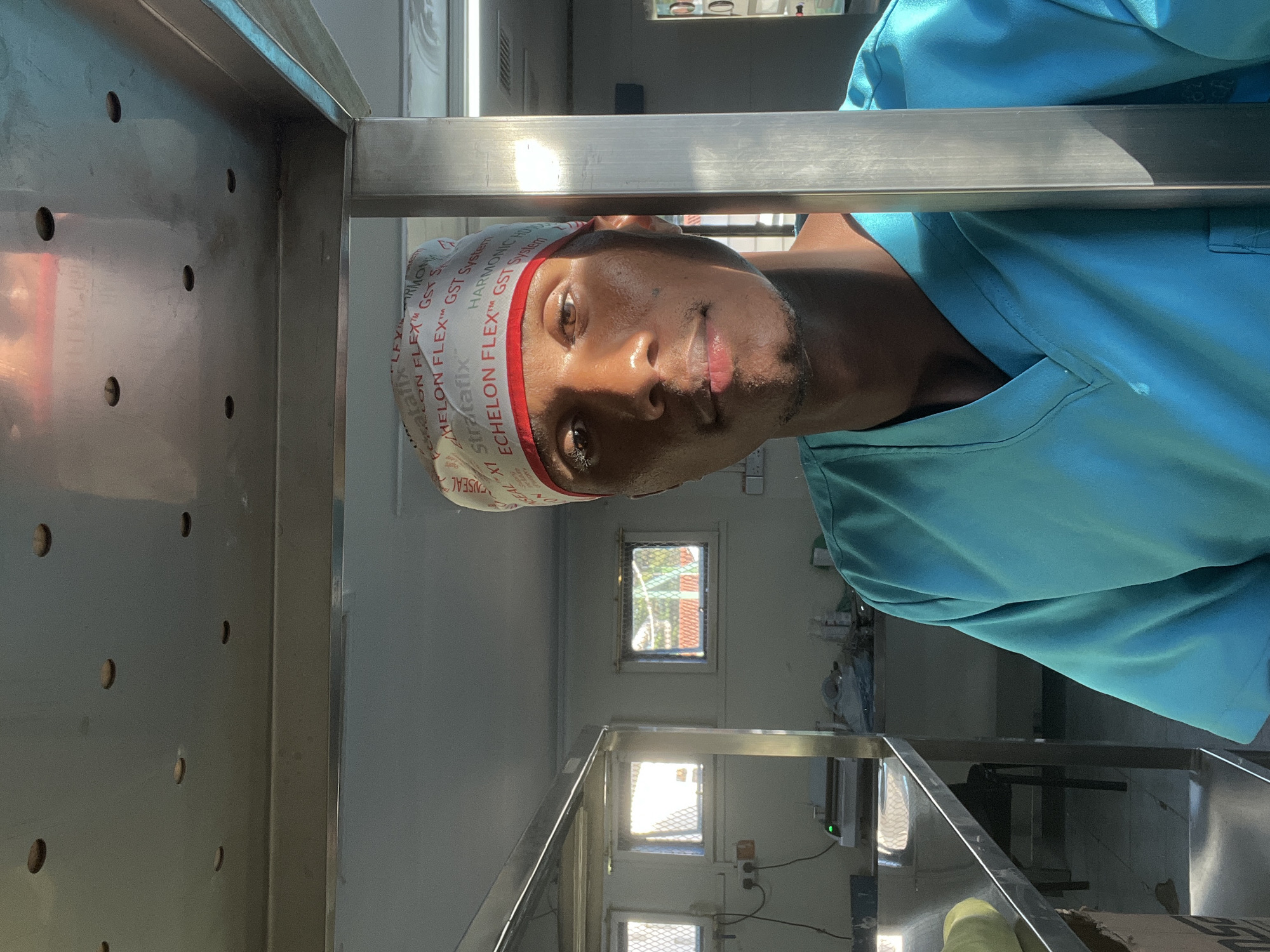 A person wearing light blue scrubs and a surgical cap with medical text, is in a medical environment. The person is centered in the image, looking at the camera. The background consists of medical equipment and storage units constructed of stainless steel. Sunlight is streaming in from windows further back in the room. The location is Nairobi, Kenya.