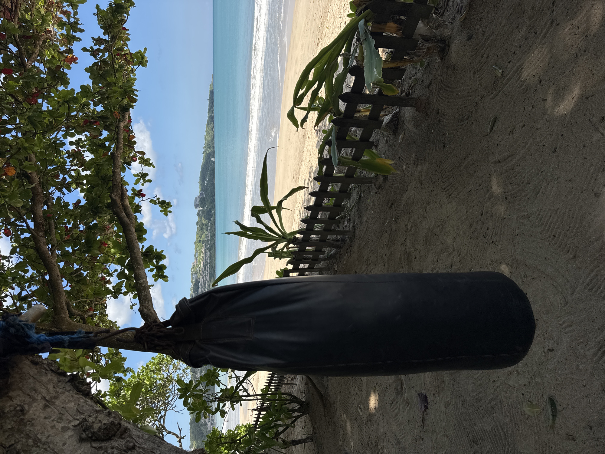 The image shows a beach scene in Kuta Selatan, Indonesia. A black punching bag hangs from a tree branch on the sandy shore, with the ocean and beach visible in the background. A dilapidated wooden fence runs parallel to the shoreline. The sky is blue with some clouds.