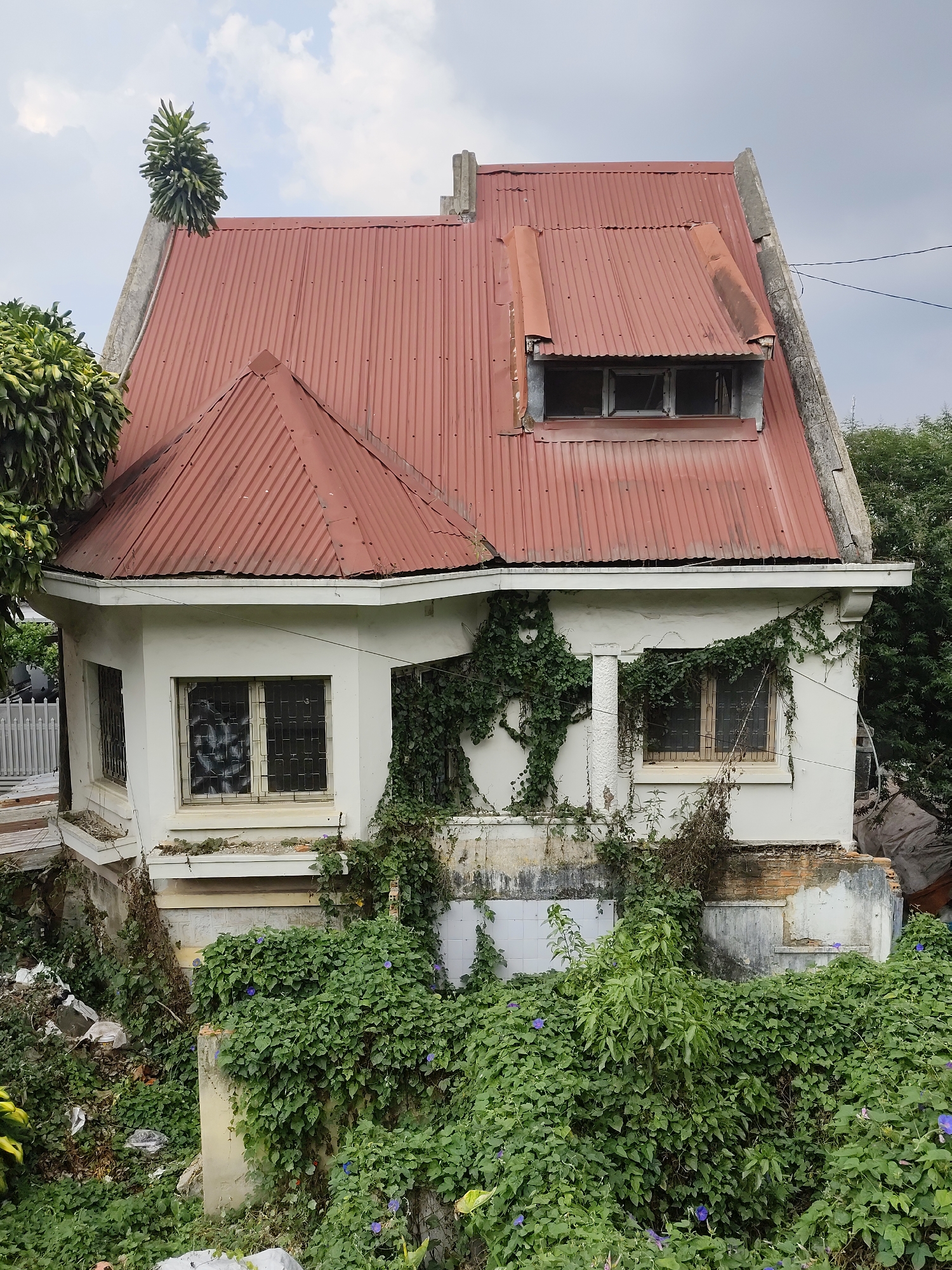 The image shows a neglected, abandoned house in Da Lat, Vietnam. The house is two stories tall with a red corrugated metal roof that has a dormer window. The exterior is white, but shows signs of decay with cracks, peeling paint, and extensive growth of ivy and other vegetation covering the walls and base. The windows are barred and appear to be covered in graffiti or obscured from the inside. The building is surrounded by overgrown plants and debris, suggesting long-term abandonment and lack of maintenance. A section of white tile appears to be on the exterior wall. There is no evidence of human activity or recent habitation in the immediate environment.