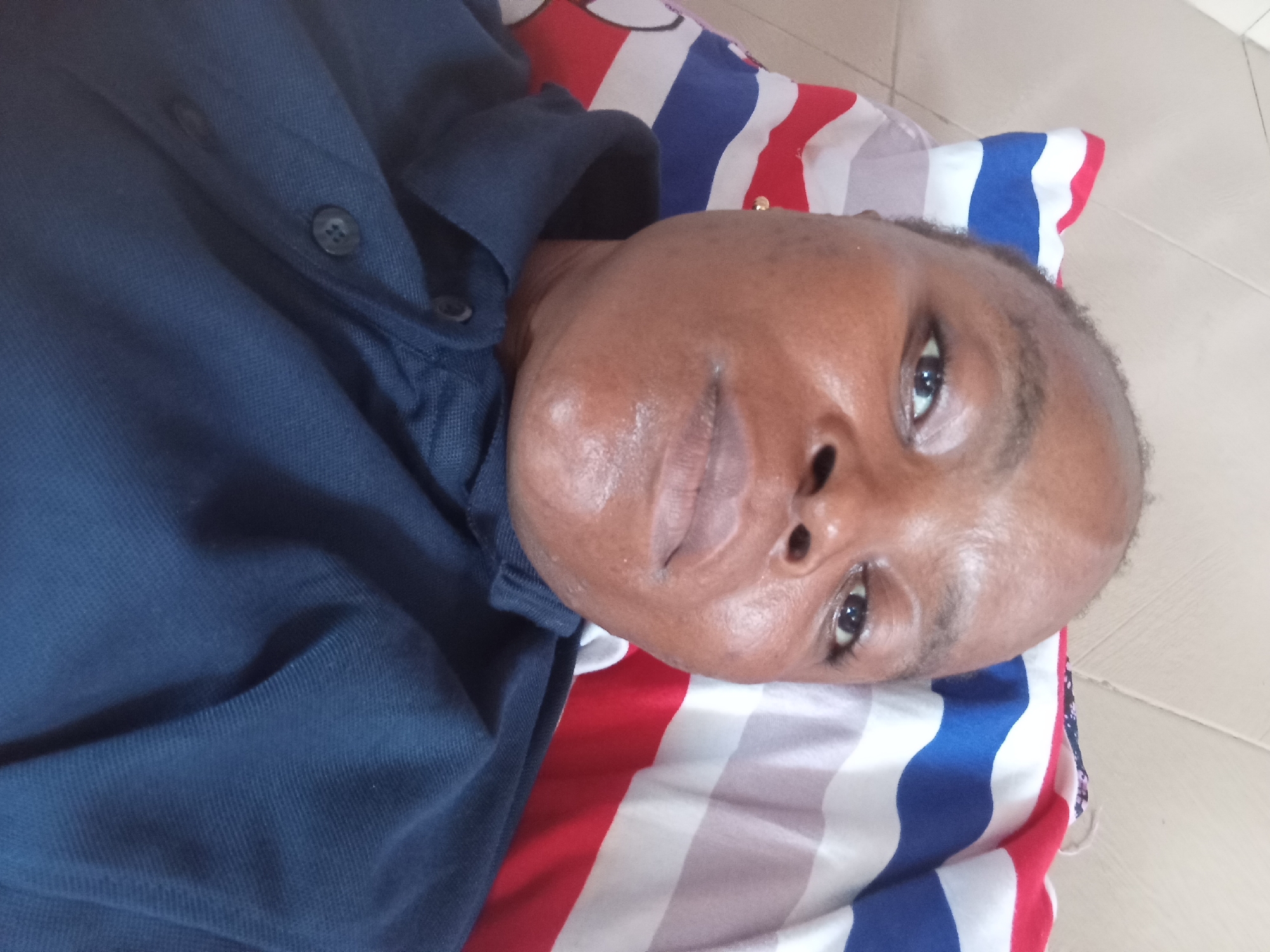 A person is lying on their back on a striped pillow. The person is wearing a dark blue collared shirt. The pillow has red, white, and blue stripes. The background appears to be a tiled floor. The image is a close-up of the person's face and upper body. The person's face is angled upwards and to the right. The person's eyes are open and looking forward. The person's lips are slightly parted. The person's skin is brown and appears to be slightly oily. The person's hair is short and dark. The person is wearing a small gold earring in their left ear. The image is taken from a slightly elevated angle, looking down at the person. The lighting is natural and appears to be coming from the right. The overall impression is of a casual, relaxed scene.
