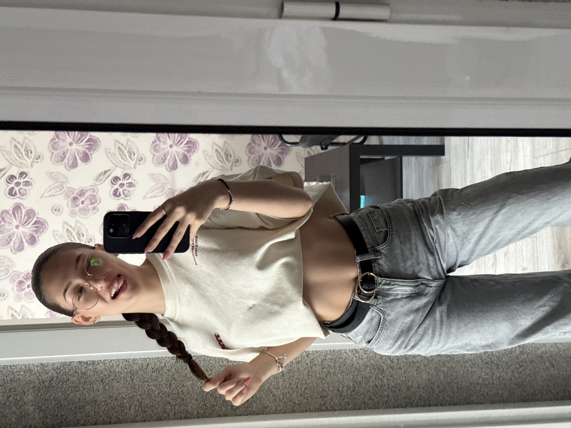 The image shows a young woman with a braid, wearing glasses, a cropped t-shirt, and jeans. She is taking a mirror selfie indoors, with a patterned wallpaper visible in the background. The scene appears to be a domestic setting, possibly a bedroom or dressing area. There is no information in the image to suggest it is located in Sofia, Bulgaria.