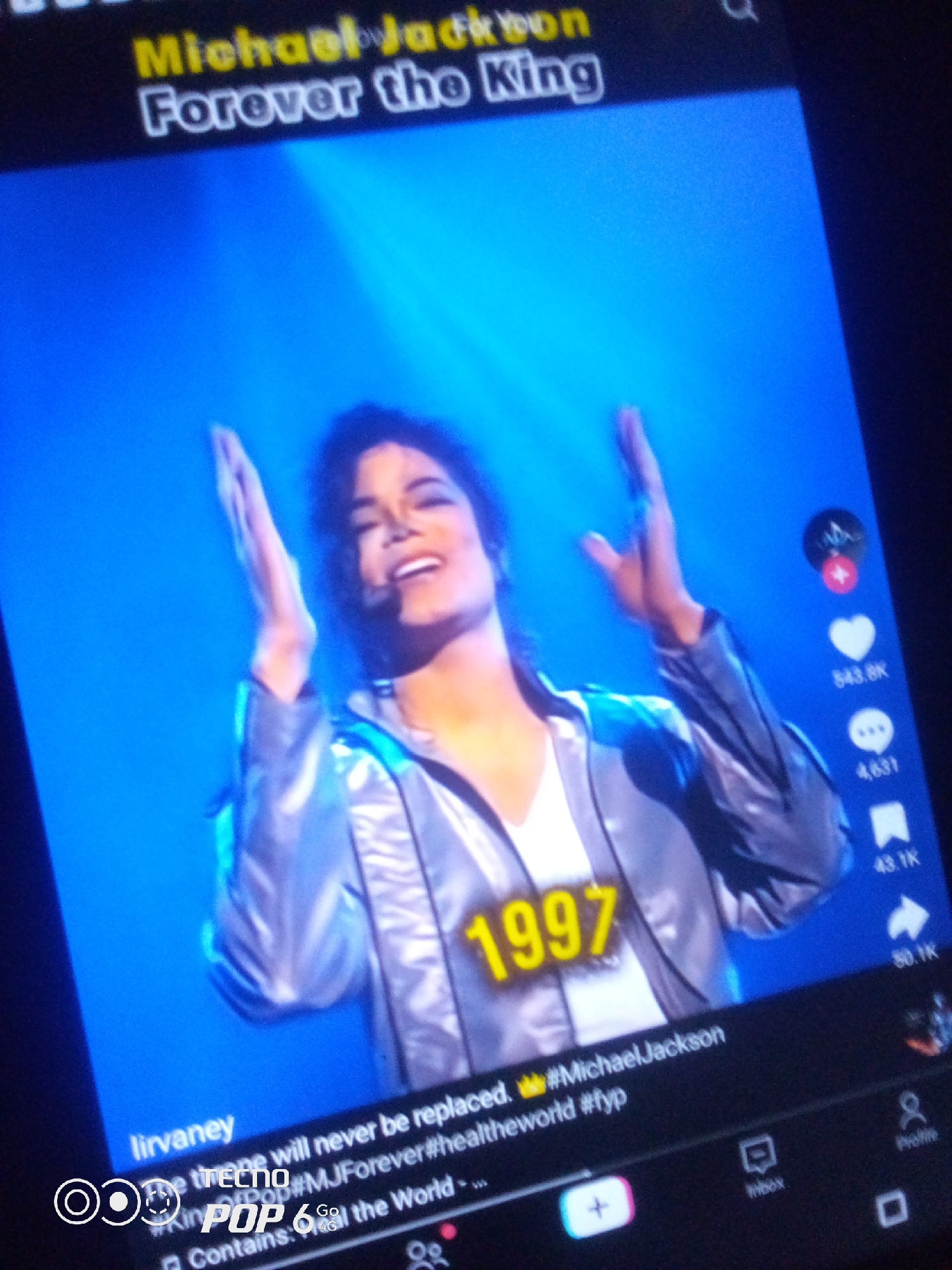 The image is a close-up, slightly blurry photograph of a screen displaying a video of Michael Jackson. He is wearing a shiny silver jacket over a white shirt and is captured mid-performance with his hands raised and a smile on his face. The background is a vibrant blue with stage lighting. Overlayed on the video are text elements, including "Michael Jackson Forever the King" at the top, and "1997" prominently displayed in yellow on Michael Jackson's jacket. Below the video, social media interface elements are visible, such as like and comment counts, as well as hashtags like #MichaelJackson and #fyp. There are also icons for inbox and profile. The screen appears to be part of a mobile device, possibly a smartphone, held by someone whose hands are not visible. The overall impression is of a social media video shared online, celebrating the artist Michael Jackson. The requested location of Howayi, Nigeria, is not depicted or implied in the image.