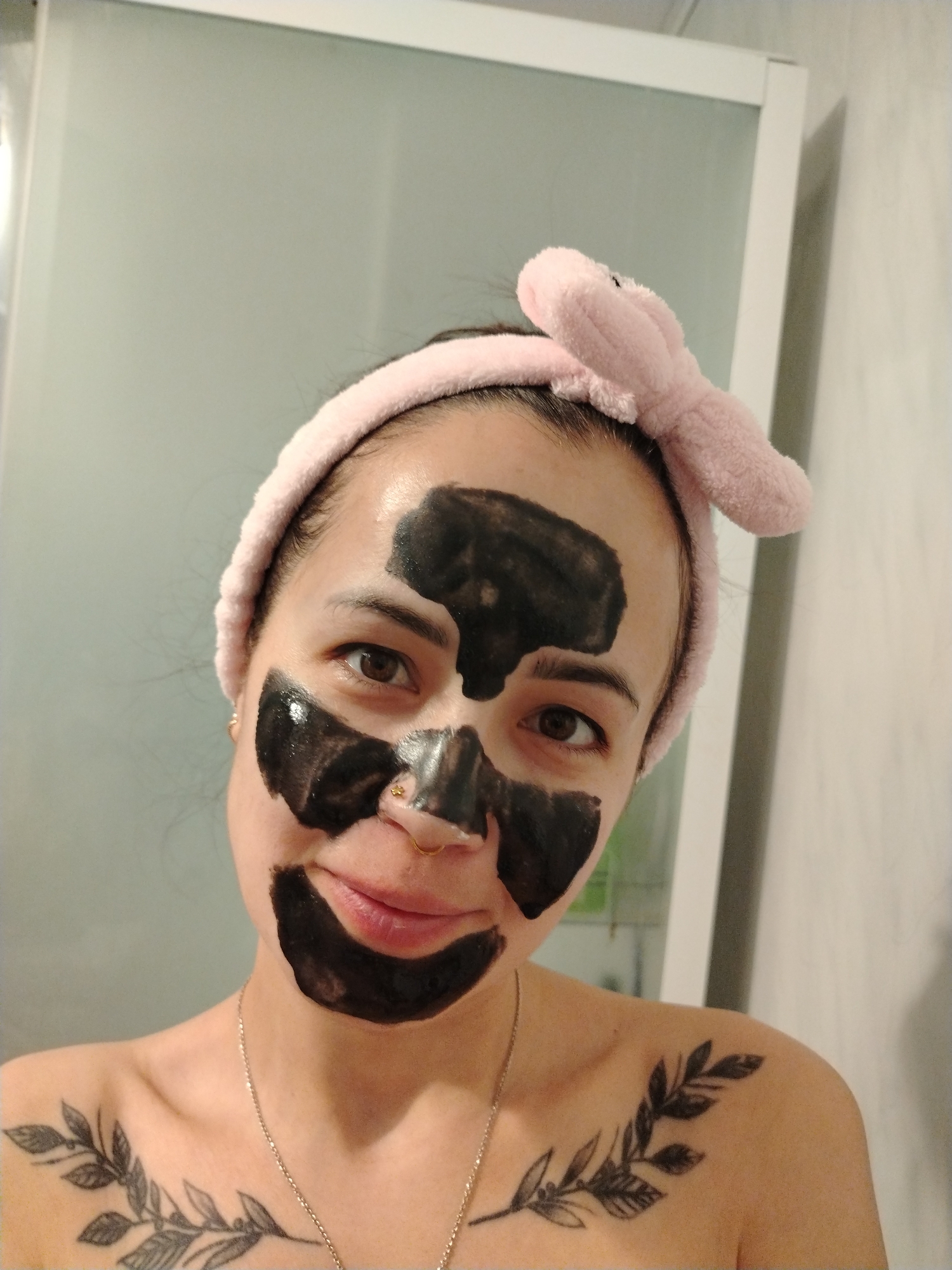 A person is posing for a selfie in a bathroom. They are wearing a pink headband with bunny ears and a black facial mask covering their forehead, nose, and mouth area. The person has a leaf-themed tattoo on their chest. They are wearing a necklace. The bathroom has white walls and a white-framed glass shower door. There is no discernible location indicating the city of Noviy, Russia.
