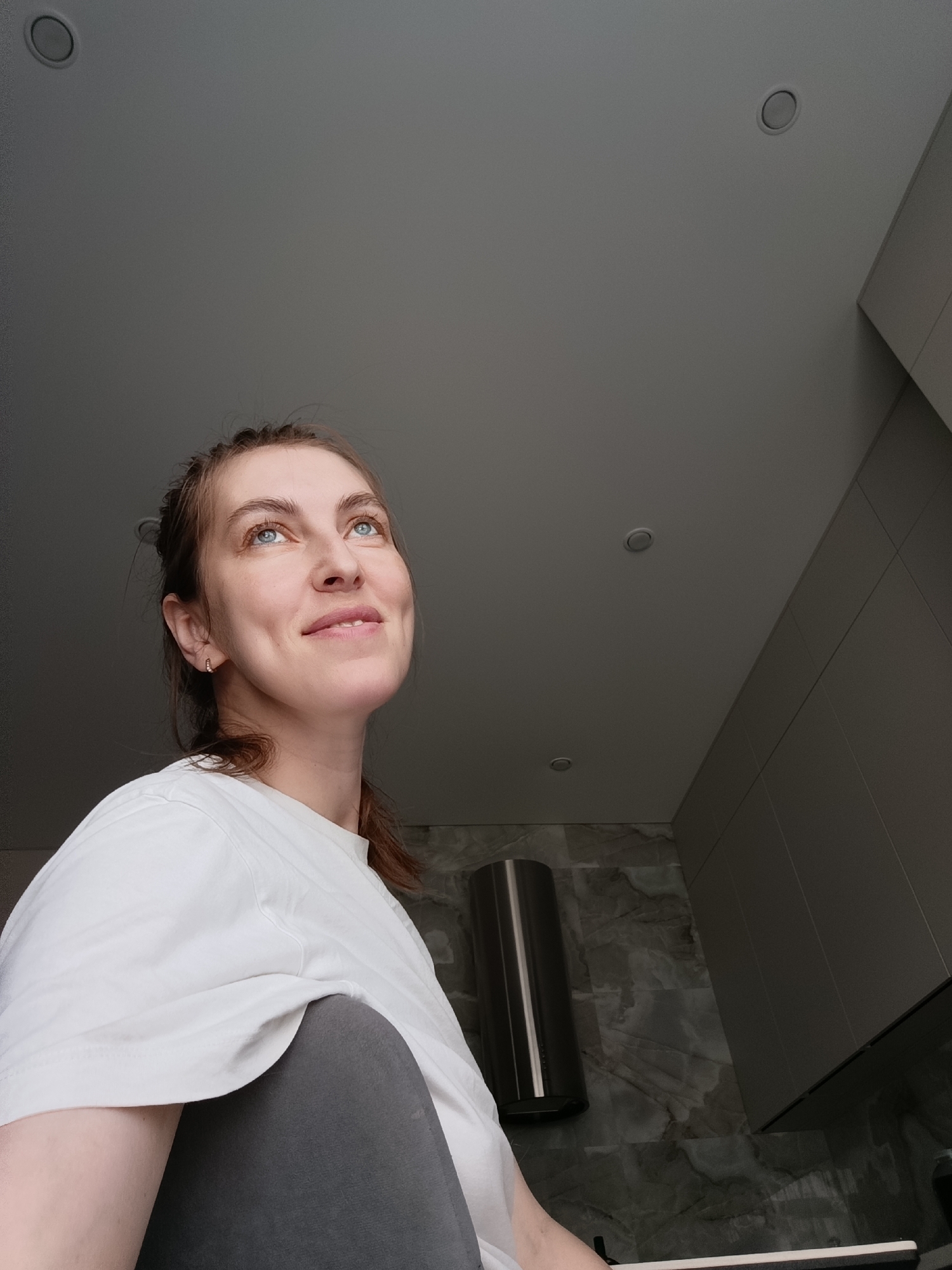 A woman with fair skin and light blue eyes is looking upwards, likely at the ceiling. Her hair is pulled back. She is wearing a white t-shirt and appears to be indoors. The ceiling is a neutral gray color and has recessed lighting. A portion of a wall with what appears to be dark gray marble-like tile is visible in the background. A stainless steel cylinder is visible on the wall. The location appears to be a bathroom or kitchen. There is no specific data in the image to definitively determine if the location is Chaikovsky, Russia.