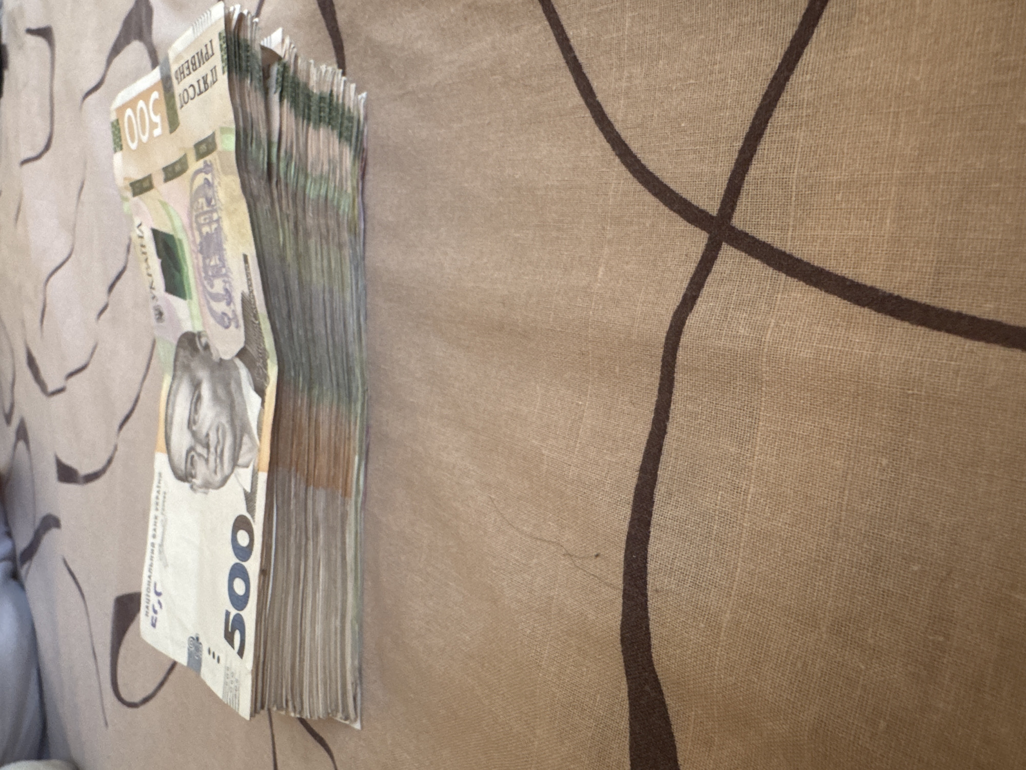 The image shows a stack of Ukrainian 500 hryvnia banknotes on a beige, textured surface. The banknotes are piled in a thick wad, with the top bill clearly visible. The top bill features a portrait of Hryhorii Skovoroda, a Ukrainian philosopher, poet, and composer. The denomination "500" is prominently displayed in blue numerals. The text "НАЦІОНАЛЬНИЙ БАНК УКРАЇНИ" (National Bank of Ukraine) is also visible. The background appears to be a fabric with a pattern of swirling dark lines. There is no indication of a specific location within the city of Mykolaiv, Ukraine.