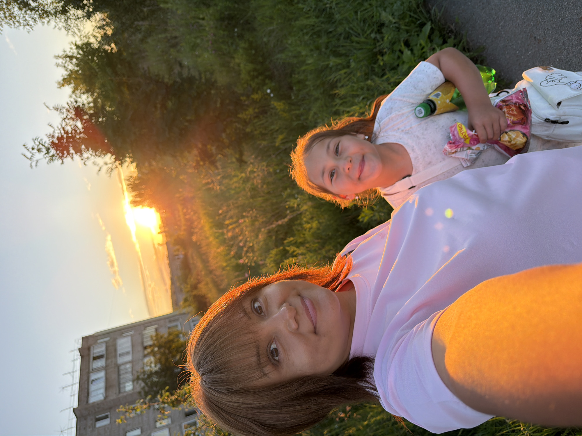 In the city of Unknown, Unknown, during sunset, a woman and a young girl are captured in a selfie. The woman, with brown hair and bangs, wears a pink t-shirt and smiles at the camera. To her right, the girl, with long brown hair, smiles and holds a snack bag and a green bottle. She is wearing a white t-shirt. A white bag with a cartoon dog print is slung over her shoulder. In the background, a tall, gray building stands to the left, while lush green trees and grass dominate the right side of the frame. The setting sun casts a warm, orange glow across the scene, illuminating the grass and the subjects.