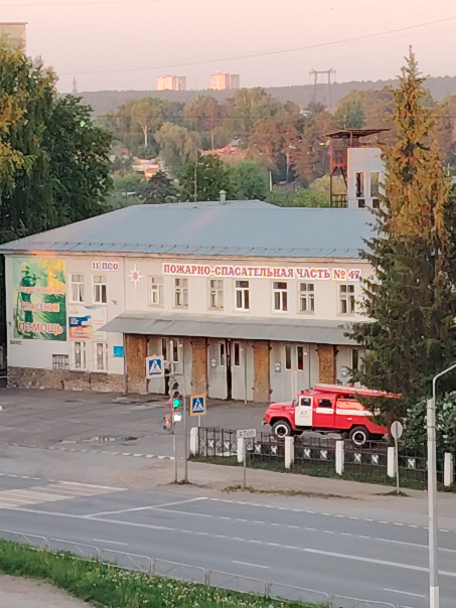 The image shows a two-story building identified as "11 PEO Fire and Rescue Unit No. 47" in Chaikovsky, Russia. The building is light-colored with a gray roof. A large banner is attached to the front of the building, along with various signs. Below the main structure is a covered area supported by pillars. A red fire truck is parked near the building. A street is in the foreground with a crosswalk and a traffic light showing a green signal. A fence runs along the side of the road. In the background, there is a cityscape of trees and buildings under a light sky. A radio tower and other structures can be seen in the distance.