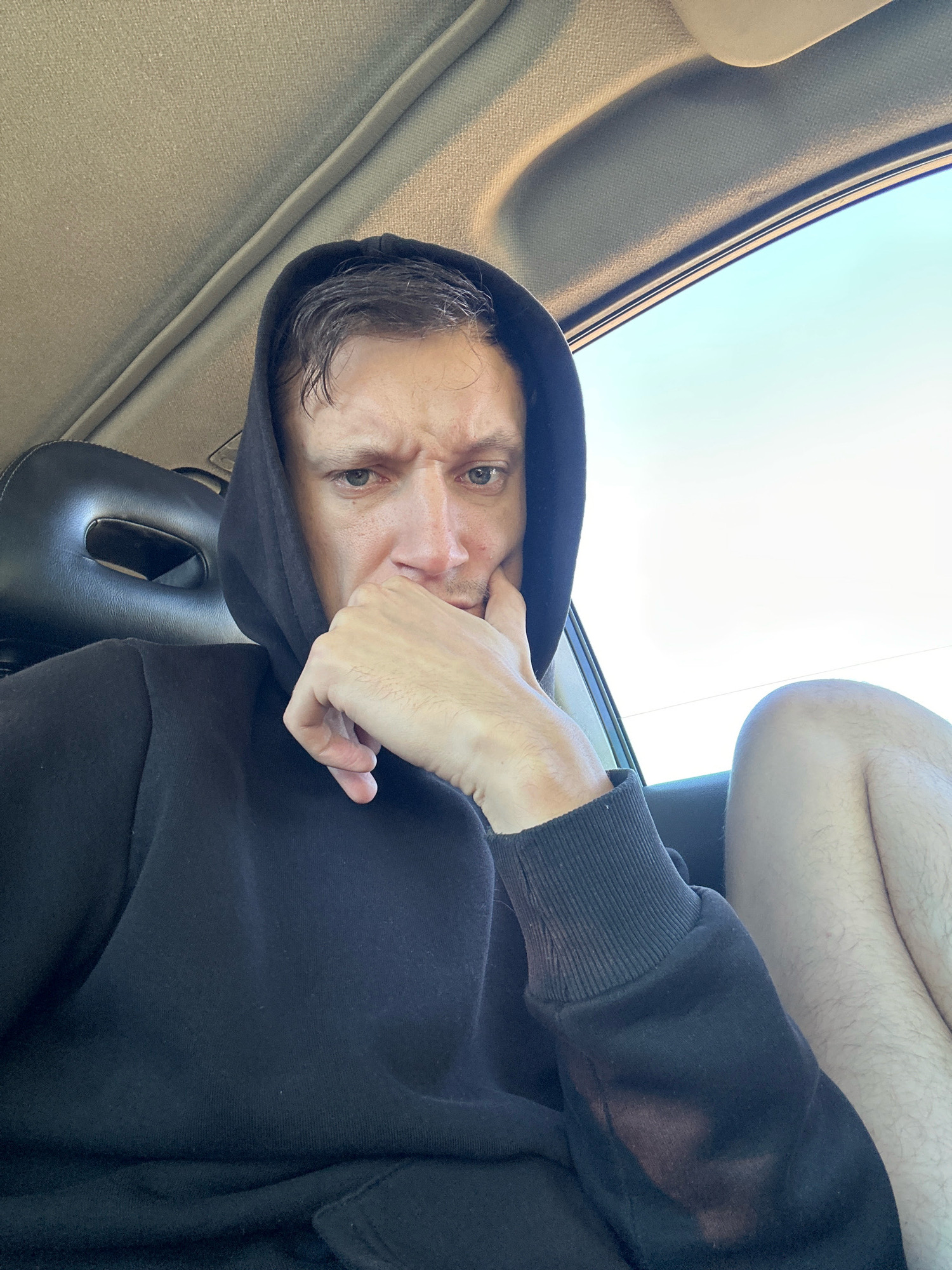 A man is sitting in a car and taking a selfie. He is wearing a black hoodie and has his hand on his chin, with a furrowed brow. The interior of the car is visible behind him, as is the bright sky outside the window. The image does not provide any information about the man's location in the city of Makiv, Ukraine.