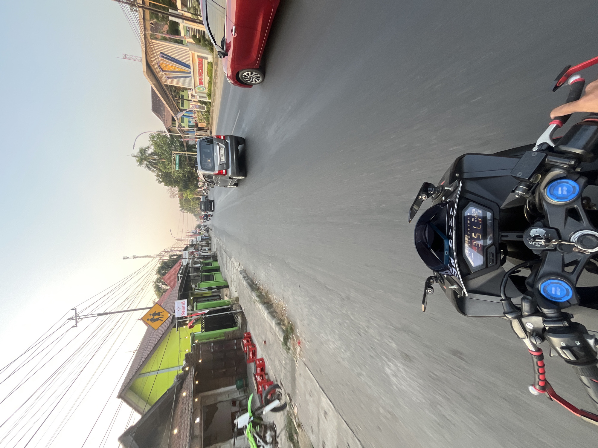 The image depicts a first-person perspective from a motorcycle traveling on a paved road in Kalinyamatan, Indonesia. The road is flanked by buildings and vegetation. On the left side, there are several small shops or eateries with outdoor seating, including red plastic chairs and tables. A green dirt bike is parked on the sidewalk. Further down the street on the left, a bright green building with a brown roof is visible, along with a yellow pedestrian crossing sign. On the right side of the road, there are more buildings, including a structure with a white facade adorned with blue and yellow accents and text that appears to be a building name. Several vehicles are on the road, including a grey minivan and a red car. The sky is clear and shows the soft light of late afternoon or early morning. The motorcycle's dashboard displays speed and other information.
