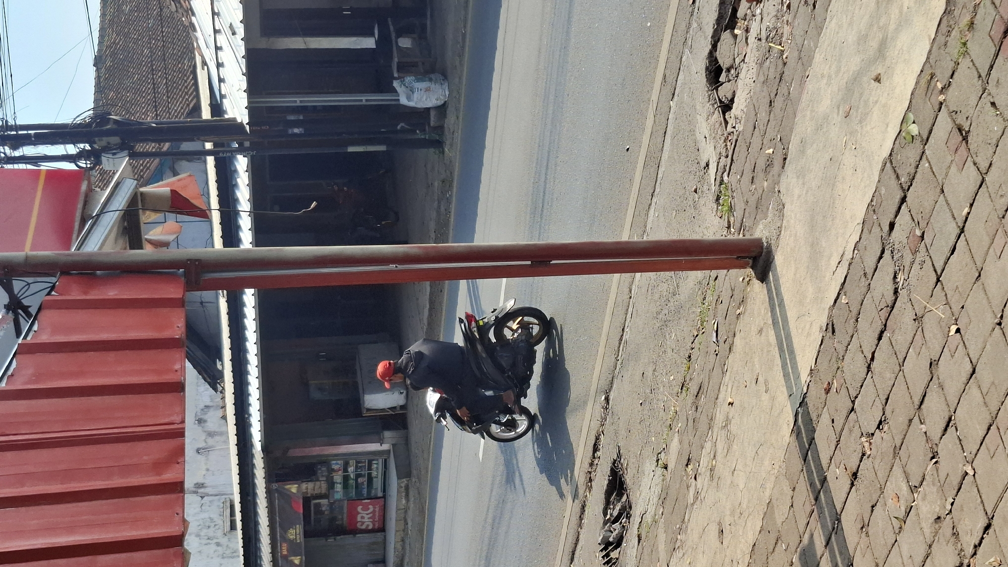 A person wearing a red cap and black jacket rides a black scooter on a paved road. The scooter has gold accents on its rear wheel. The person is in motion, casting a shadow on the road. The road is bordered by a sidewalk made of interlocking bricks and concrete. There are shops in the background with awnings and a red metal structure on the left. Utility poles with wires are visible in the background. The scene appears to be in a commercial or residential area on a sunny day. The location is identified as Cilaku, Indonesia.