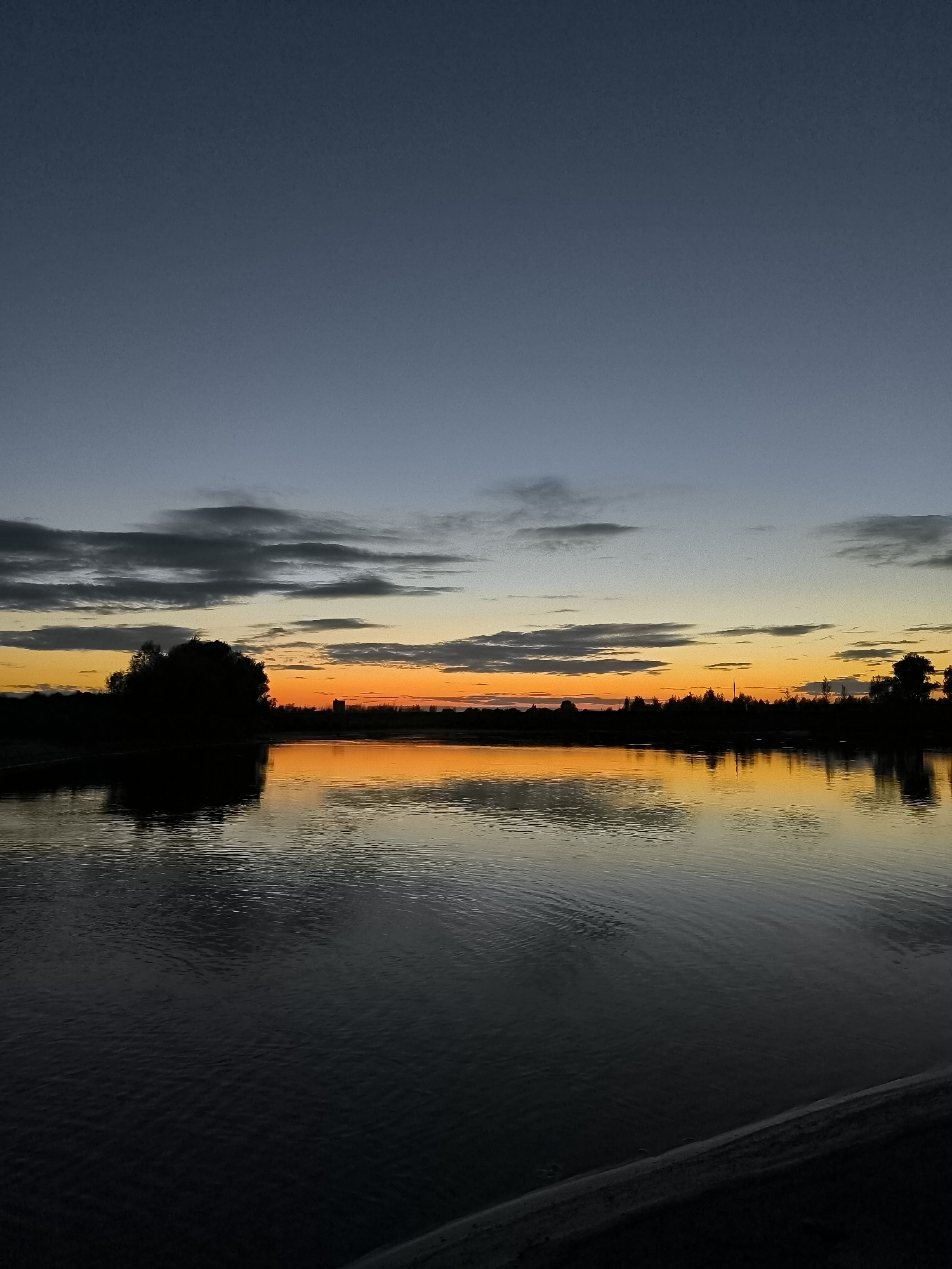 The image depicts a tranquil natural scene at dusk, likely in Zagotzerno, Russia. A body of water, possibly a lake or river, dominates the foreground and midground, reflecting the twilight sky and the silhouetted landscape. The water's surface is calm, with gentle ripples creating subtle patterns.

The horizon line is defined by a dark silhouette of trees and vegetation, with a prominent, large, bushy tree on the left side. In the distance, on the right side of the silhouette, a few faint, vertical structures, possibly utility poles or buildings, are visible.

Above the treeline, the sky displays a gradient of colors characteristic of sunset. A vibrant band of orange and yellow hues is present near the horizon, transitioning into a dusky blue towards the top of the frame. Scattered clouds, appearing in shades of gray, are visible in the upper portion of the sky. The overall atmosphere is serene and quiet, with no human presence or activity evident. The scene is captured from a low vantage point, possibly from a shoreline or a low embankment, with a dark, indistinct element in the bottom right corner that might be a railing or edge of a structure.