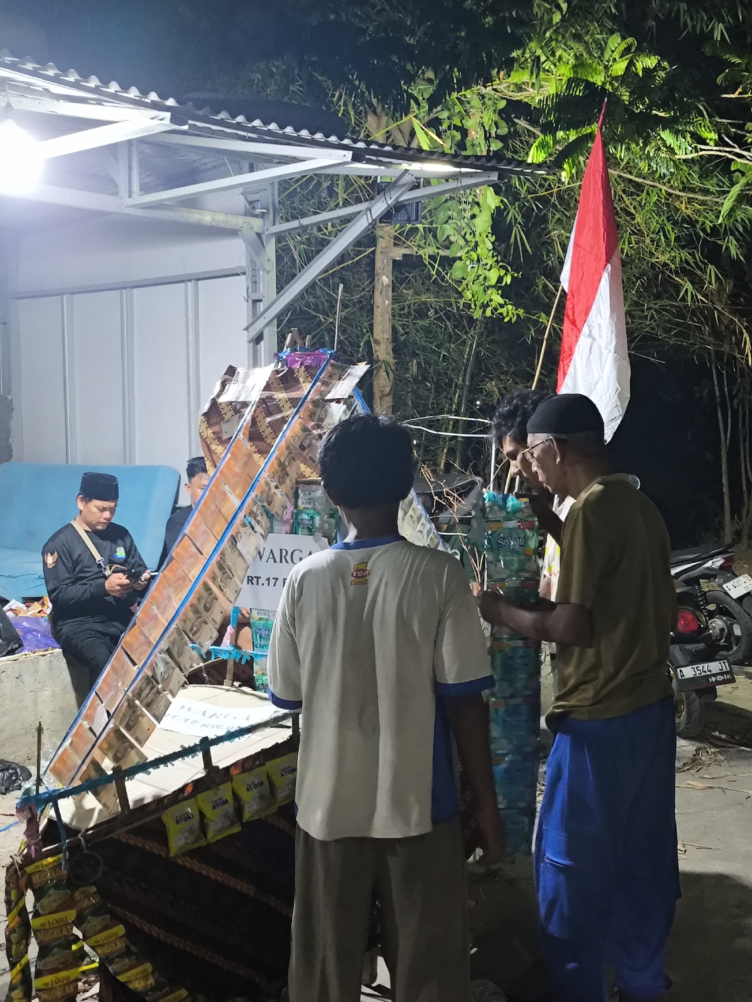 In the city of Cilegon, Indonesia, at night, a group of men are gathered around a display. The scene is illuminated by artificial lights, creating a somewhat theatrical atmosphere. In the foreground, three men are visible. The man on the far right, wearing a dark green t-shirt and blue pants, is looking down and appears to be holding something in his hands. To his left, a man in a white and blue shirt faces away from the camera, also looking towards the display. Another man in a black shirt and black cap is seated to the left, looking at his phone.
The central focus of the image is a tall, multi-tiered display, possibly a form of stage or decoration, constructed from cardboard and other materials. It is adorned with what appear to be snacks or small packets, some with yellow packaging and text. A red and white Indonesian flag is prominently displayed on a pole behind the structure. To the right of the display, a motorcycle is parked, with its license plate clearly visible as "A 3544 JT". The background consists of dense foliage, suggesting a natural or semi-natural setting. The overall environment appears to be outdoors, possibly in a yard or public space.
