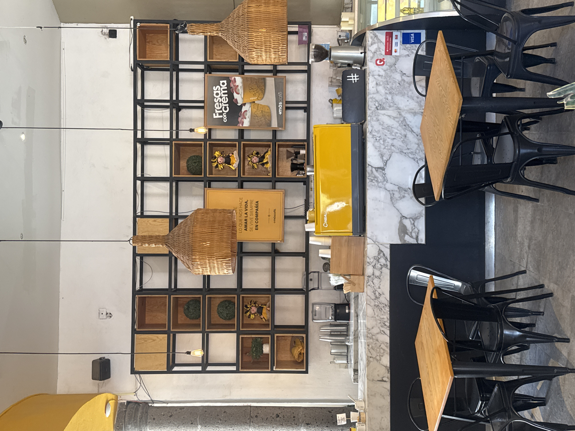 The image shows the interior of a cafe or restaurant, most likely in Bernal, Mexico. The scene features a black metal shelving unit with various items displayed inside, including wooden boxes, decorative balls, dolls, and signage. Notable signs promote "Fresas con Crema" and a tagline about "Amar la Vida" (Love Life). A yellow coffee machine is positioned next to the shelving unit. The cafe also has marble countertops and black chairs arranged around small wooden tables. The image includes light fixtures hanging from the ceiling. The environment is well-lit. There are no people visible in the frame.