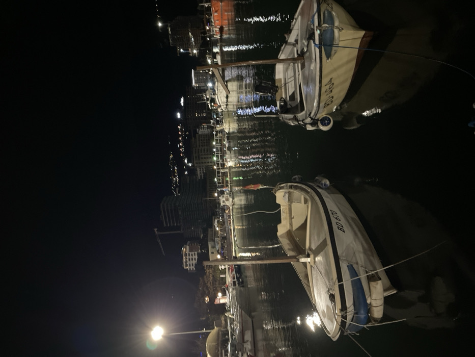 Two small boats are docked in a harbor at night. Buildings line the waterfront, their lights reflecting in the calm water. The boats are white with dark trim, and identifiable by markings "BD 670" and "BD 464." A single street light illuminates the scene in the foreground. The overall atmosphere is peaceful and quiet.