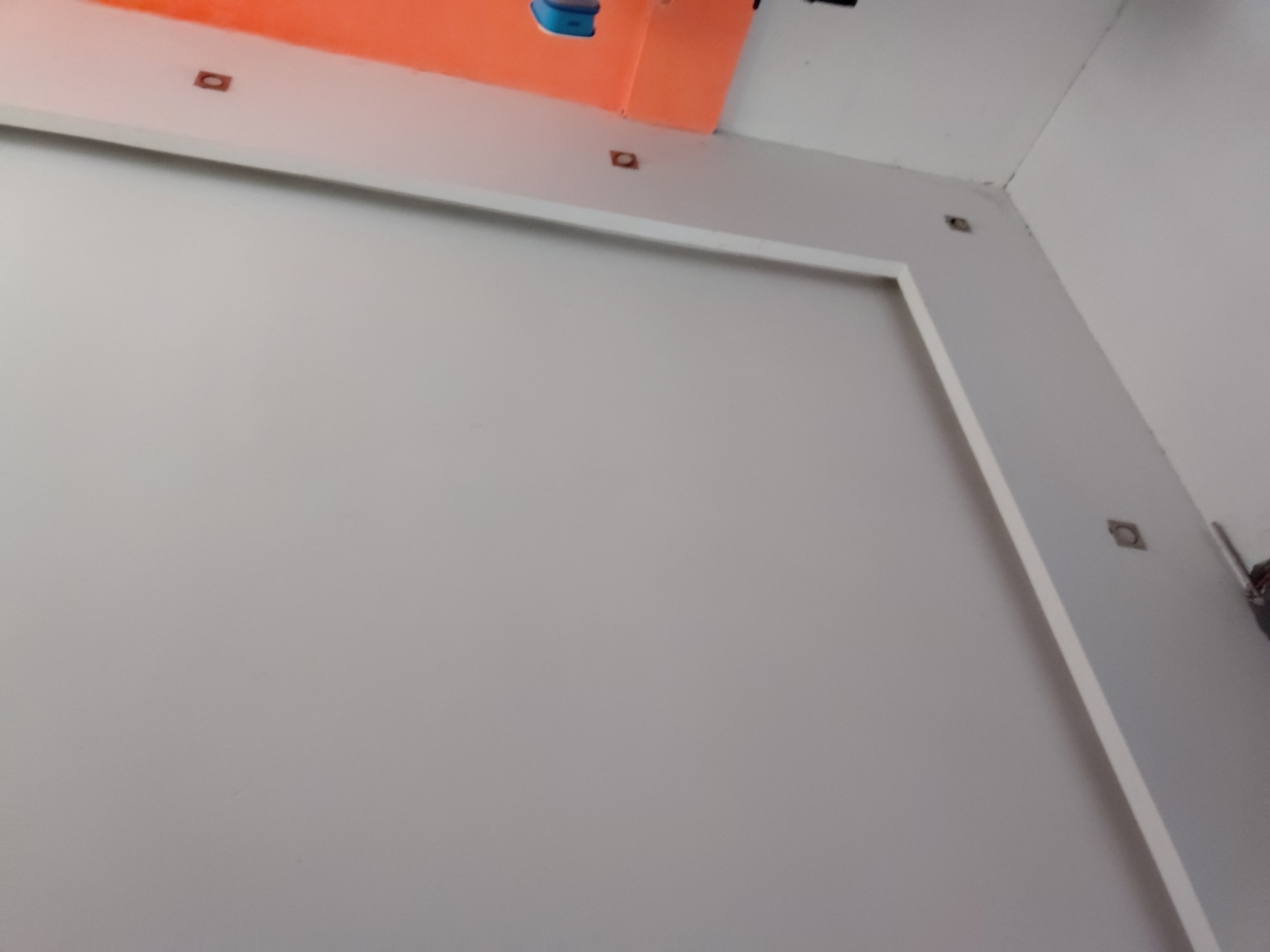 The image displays an indoor scene, likely a room under construction or renovation, located in Santa Ana, Venezuela. No people are visible.
The primary subject is a large, light gray wall that occupies most of the frame. A white, rectangular trim or frame outlines a section of this gray wall, creating a distinct border. Above this trim, the ceiling or an upper wall section is painted a vibrant orange color, extending across the top of the image from left to right.
Several small, square-shaped anchors or mounting points are visible. Two of these are embedded in the white trim near the orange upper section on the left side, and another is present towards the top right corner where the white trim meets the orange surface. A fourth identical anchor is visible on the right-hand wall, just below the ceiling line.
In the top center, on the orange surface, a small blue object, possibly a cap or part of an electrical fixture, is discernible. Toward the top right corner, some dark, indistinct objects, possibly wires, pipes, or components of a fixture, are visible against the white wall and ceiling. The overall impression is one of an unfinished or recently installed interior space, characterized by clean lines and distinct color blocking. No visible text is present.