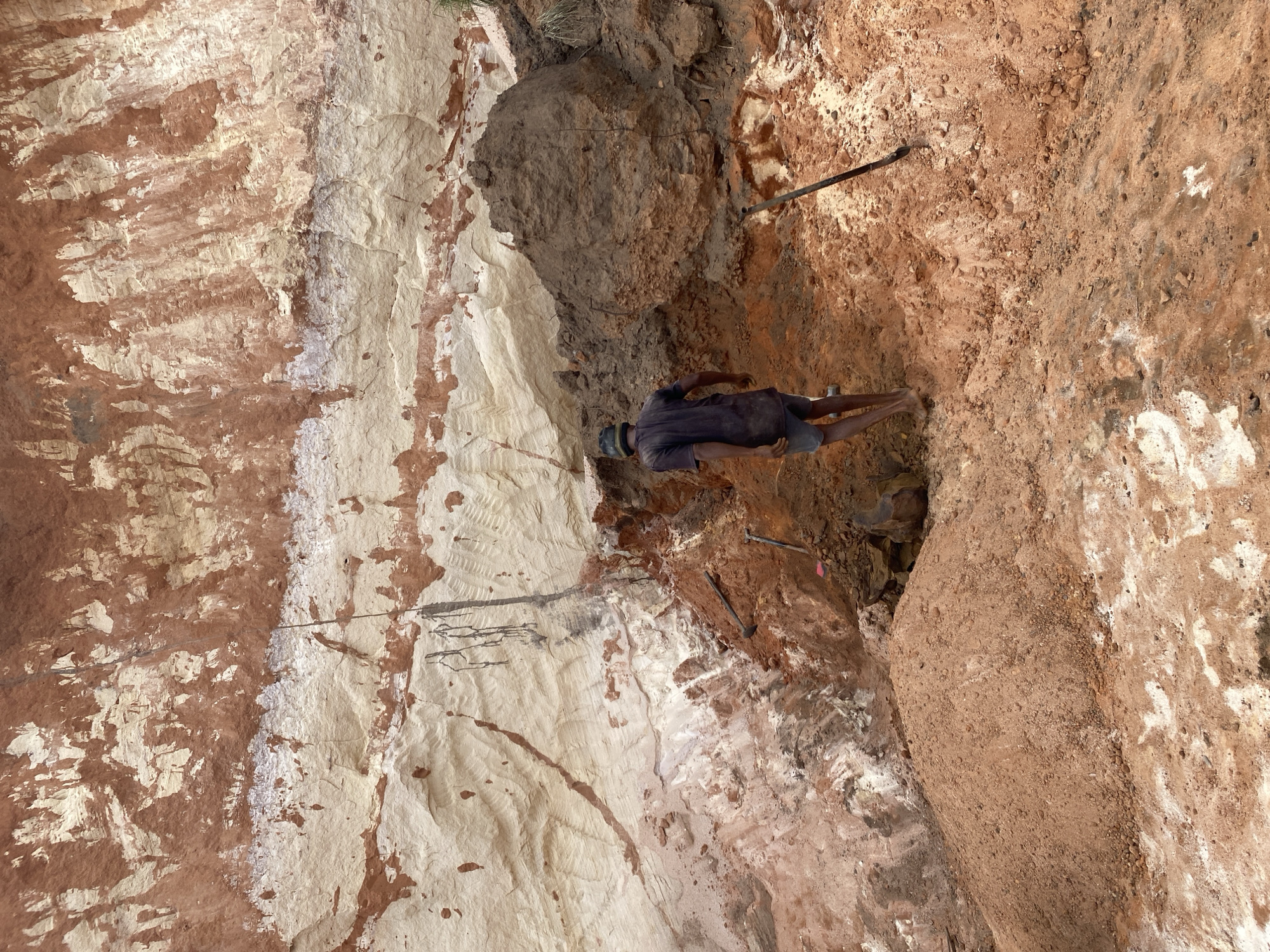 A man in a dark shirt and shorts is digging in a deep pit in the city of Amoso, Nigeria. The pit is surrounded by layers of reddish-brown and beige soil. Two long metal rods are embedded in the soil near the man. A larger pile of dark brown soil is visible above the man.