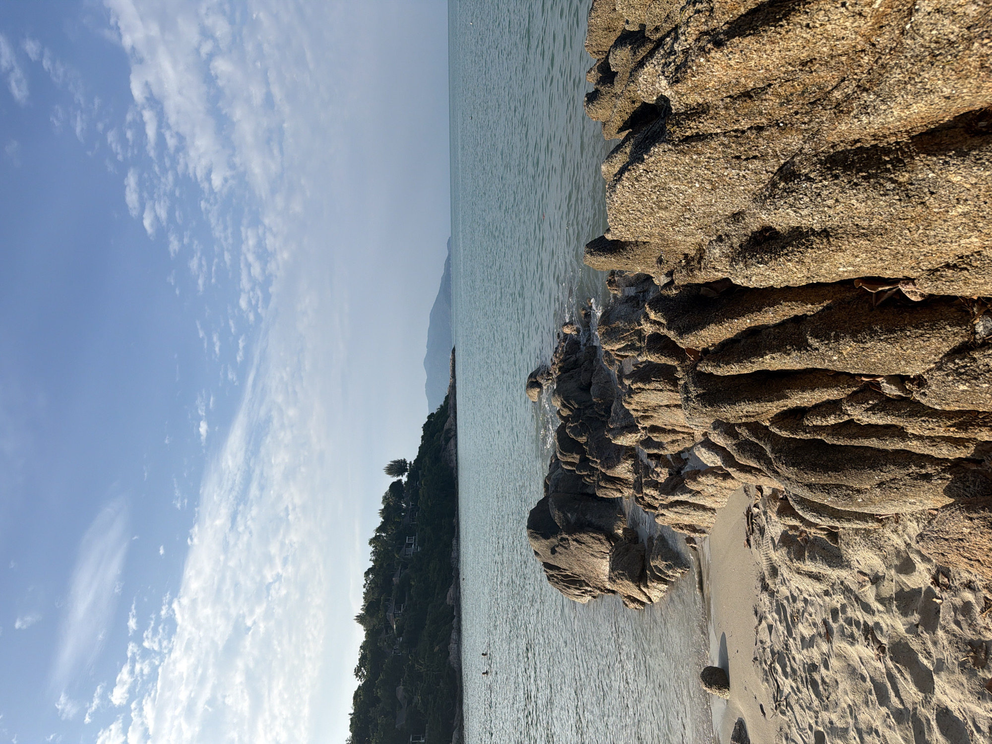 The image depicts a scenic coastal view on the island of Ko Samui, Thailand. In the foreground, rough, textured rocks are prominently featured, with their surfaces catching the sunlight. To the left, a sandy beach meets the calm, turquoise ocean. A few scattered leaves are visible on the sand. In the middle ground, the ocean stretches out, and two individuals are seen swimming or wading in the water. Further in the distance, a lush, green hillside is covered with trees and dotted with buildings, suggesting a residential or resort area. The sky above is a clear blue, adorned with scattered white clouds, indicating a bright and pleasant day. The horizon line is visible where the water meets the sky, with faint mountain ranges visible in the far distance. The overall impression is one of a tranquil tropical beach environment.