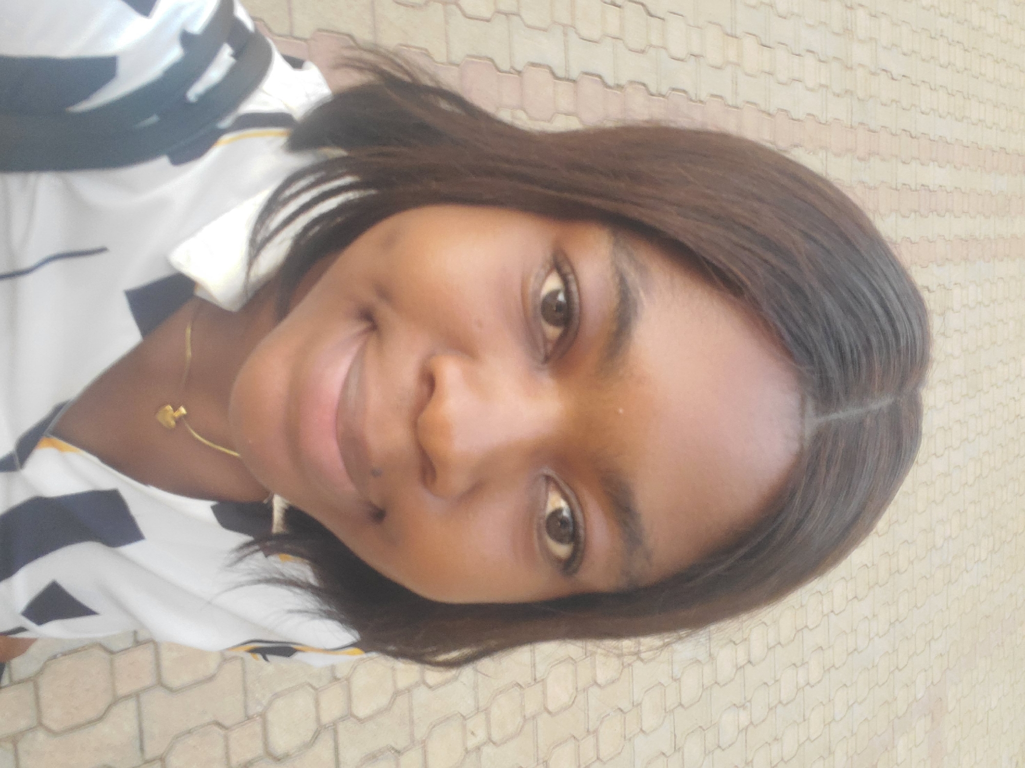 A woman with brown hair and brown eyes is smiling at the camera. She is wearing a white shirt with black stripes and a gold necklace. The background is a paved walkway. The location appears to be an outdoor area in Barden Yana, Nigeria.