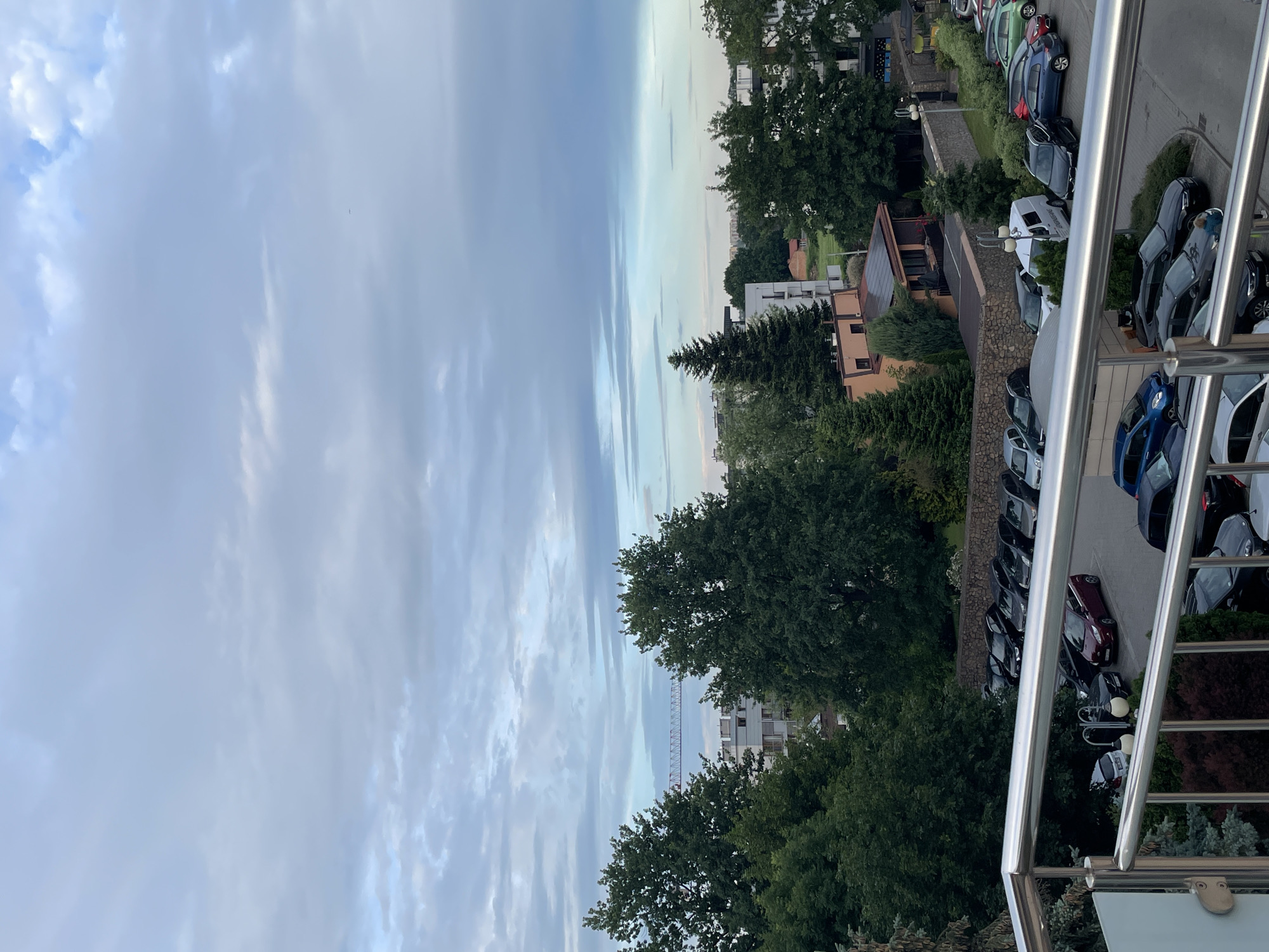 The image is a vertical, full shot of an outdoor scene. The upper half of the image is dominated by a cloudy sky with shades of blue and gray. In the middle ground, there are several large, mature trees with dense green foliage. Behind the trees, buildings and a construction crane are visible. The lower half of the image shows a parking lot filled with various cars, including sedans, SUVs, and a white van. A silver railing from a balcony or observation deck is in the foreground, partially obscuring the view of the parking lot. The ground in the parking lot appears to be paved. The environment suggests a residential or mixed-use urban area during daylight hours, possibly late afternoon or early evening given the lighting.
