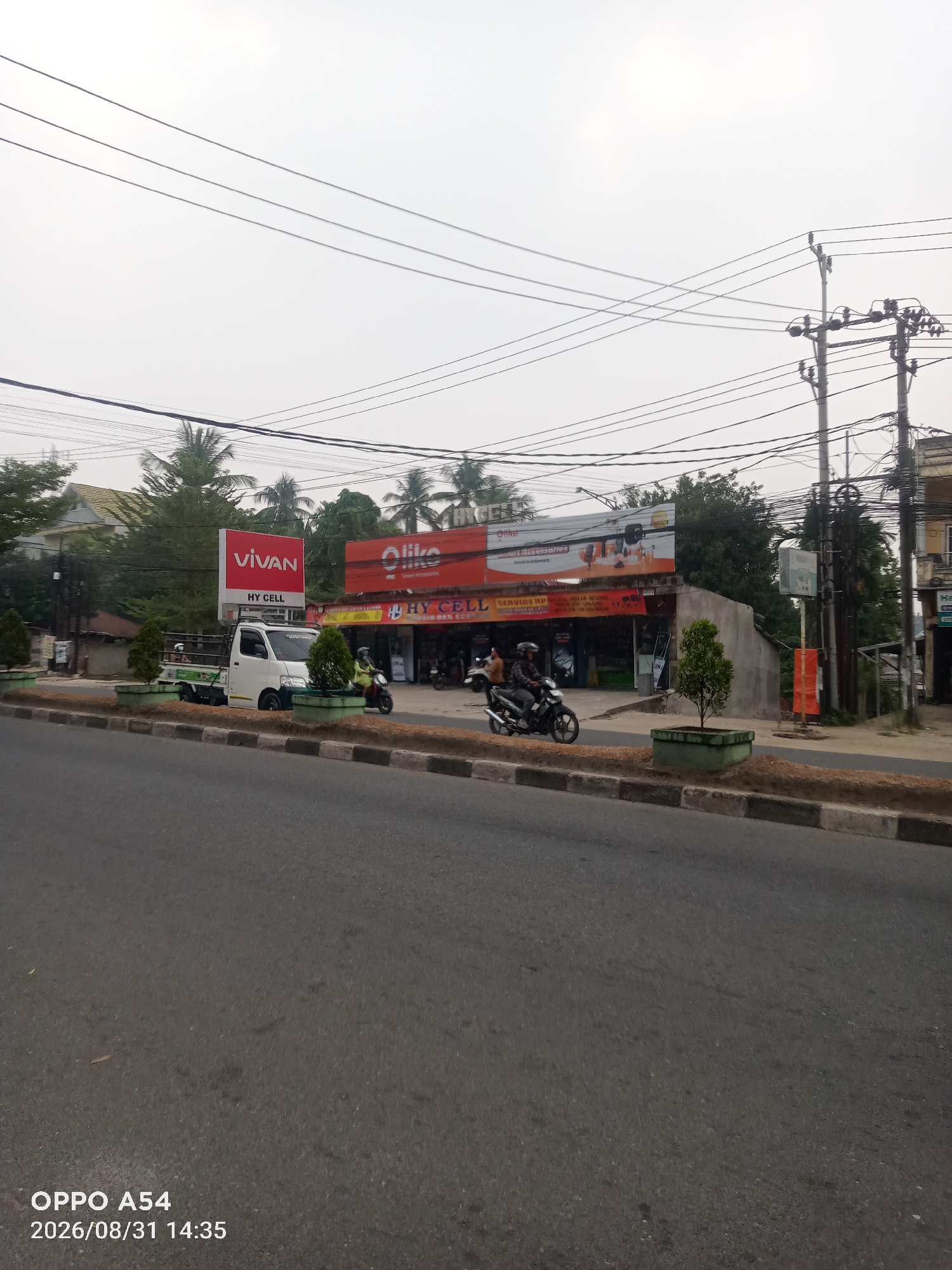 The image depicts a street scene in Jambi, Indonesia, under an overcast sky. In the foreground, a paved road runs diagonally across the frame, bordered by a raised concrete curb with black and white markings. Beyond the curb, a strip of land with scattered green planters and small trees separates the road from the sidewalk.
The middle ground features several commercial establishments. A prominent red billboard with the white "VIVAN" logo and "HY CELL" text is mounted above a white truck parked on the side of the road. Next to it, another red and white billboard displays the "Olike" logo and "Smart Accessories" text. Below these signs are storefronts, including one labeled "HY CELL SERVICE HP," with displays of electronic goods visible through the windows. Two motorcycles are present: one is parked, and another is being ridden by a person wearing a helmet and a yellow shirt, moving away from the camera.
In the background, a dense arrangement of power lines and poles dominates the upper portion of the scene, crisscrossing against the pale sky. Trees and buildings are visible behind the commercial establishments. The overall impression is that of a busy commercial area on a typically cloudy day. A timestamp in the lower-left corner reads "OPPO A54 2026/08/31 14:35".