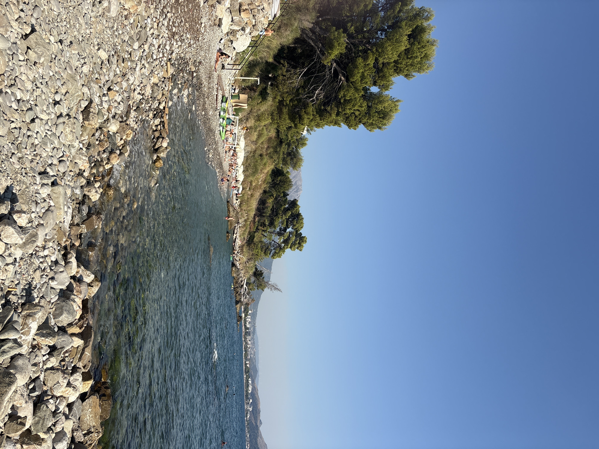 This image depicts a sunny day at a rocky beach in the city of Bar, Montenegro. The foreground is dominated by a dense arrangement of smooth, grey and white pebbles of varying sizes. To the left, a stone embankment with a black metal railing forms stairs leading up to a green, vegetated slope dotted with pine trees. Further along the shore, several people are visible lounging on the beach or in the shallow water. They are spread out, with some sitting on towels or sun loungers under white umbrellas, while others are in the sea. In the mid-ground, the clear, blue-green water gently laps the shore, revealing the rocky seabed. A few inflatable kayaks or paddleboards are present on the beach. In the background, a town with white buildings stretches along the coastline, nestled between rolling green hills and mountains under a clear blue sky. The overall scene conveys a relaxed atmosphere of a summer day at a coastal resort.