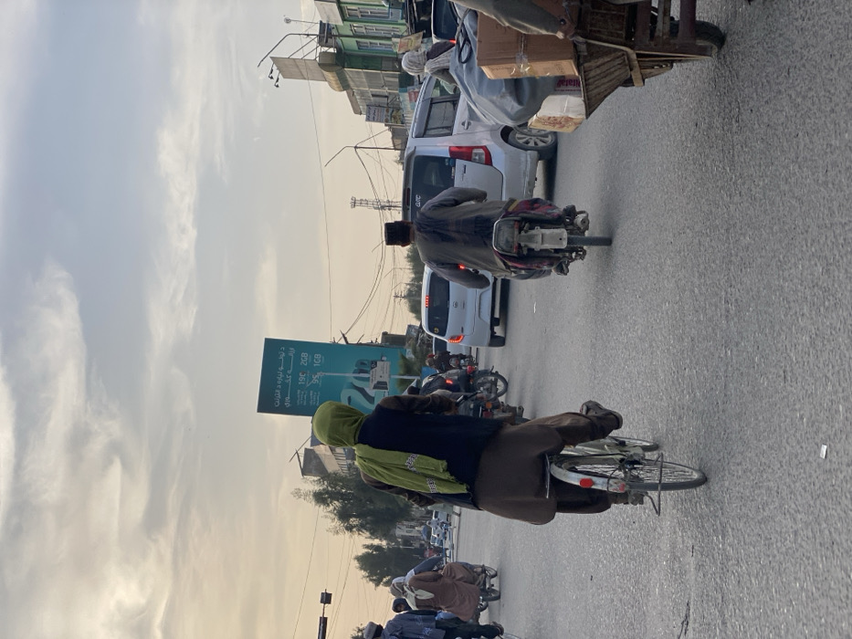 This image captures a street scene in Kandahar, Afghanistan, likely during late afternoon or early evening. The view is from behind multiple individuals moving forward on a paved, multi-lane road. The sky above is largely overcast with scattered cirrus clouds, showing faint warm hues near the horizon, indicative of a low sun angle.
In the immediate foreground, a person on a bicycle occupies the left lane, pedaling away from the camera. This individual wears a dark vest over a long-sleeved garment, loose brown trousers (shalwar), and a bright green scarf covering their head and draped over their back. To the right, partially cut off by the frame, is a handcart loaded with cardboard boxes and a dark, bundled item, being pushed by an unseen individual whose lower body and left arm are visible.
In the mid-ground, a person on a motorcycle, also facing away, drives in the right lane, wearing dark clothing and a dark cap. Behind this motorcycle, a silver compact hatchback car (possibly a Suzuki Wagon R) and a lighter grey sedan are visible in traffic. Further back on the left, another person on a motorcycle is seen, and multiple other individuals are on bicycles or motorcycles, all moving in the same direction.
The background features a busy urban environment. A prominent teal-colored billboard stands on the right, displaying text in Dari or Pashto alongside numbers such as "19C 2GB," "9C 1GB," and "20," suggesting a mobile data advertisement. Various multi-story buildings, some appearing commercial with shopfronts, line both sides of the street. Overhead utility poles and a dense network of power lines crisscross the sky. A traffic light pole is visible on the far left. No direct interactions between individuals are discernible; the scene depicts general traffic movement.