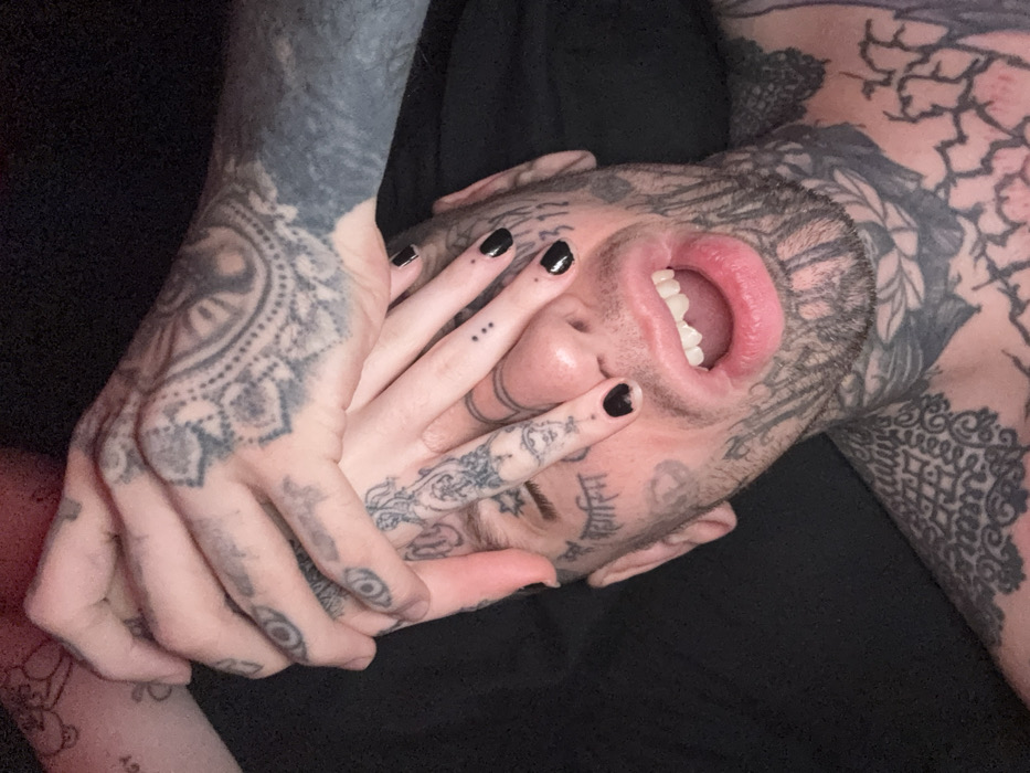 A person with extensive tattoos on their face, neck, chest, and arms lies with their mouth open and teeth visible. One hand, also heavily tattooed, is near their face. Another hand, with black nail polish and small tattoos, covers part of their face. The skin is pale, and the background is dark, creating a high-contrast image.